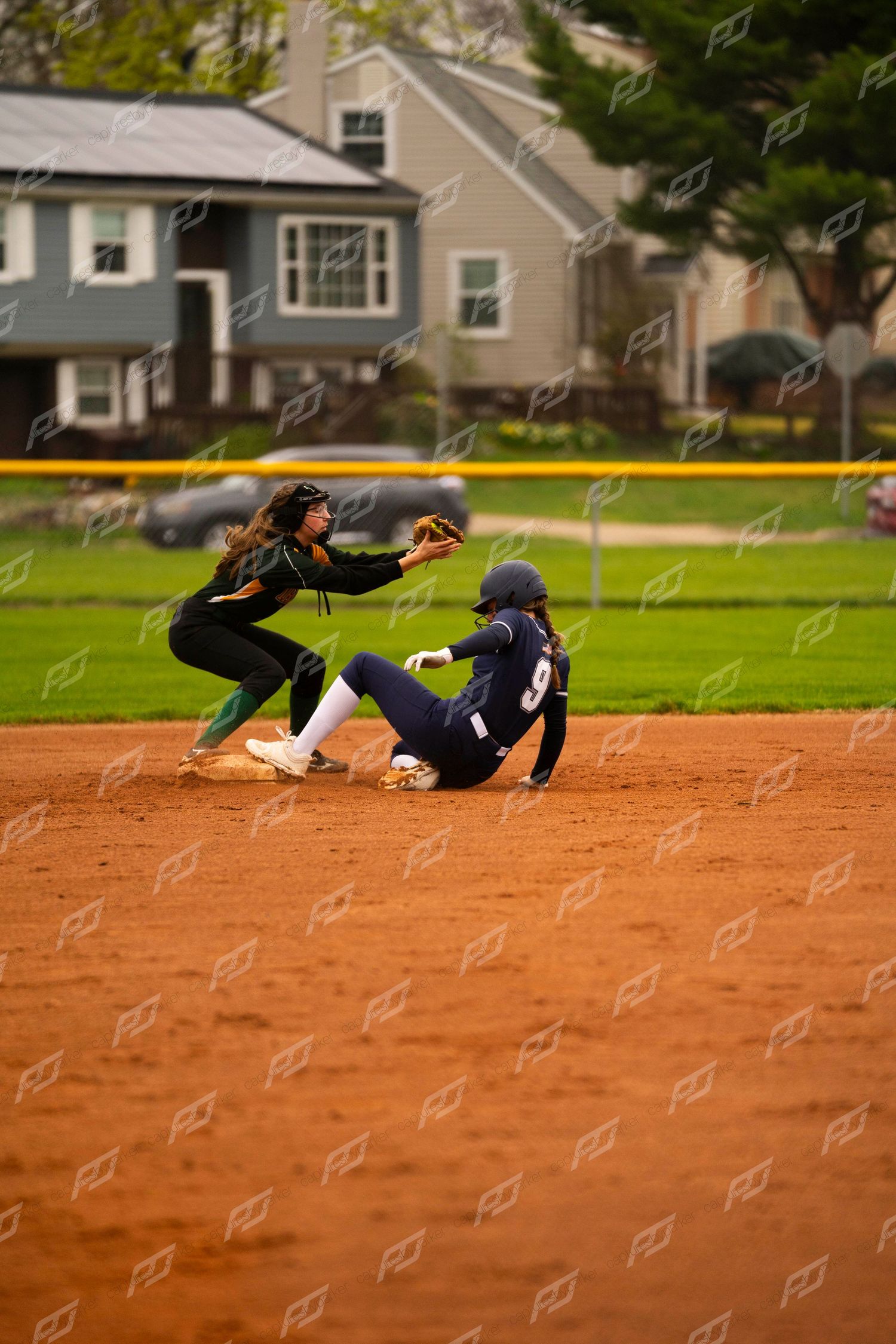 Softball player in navy uniform slides into second base as fielder in green attempts tag.