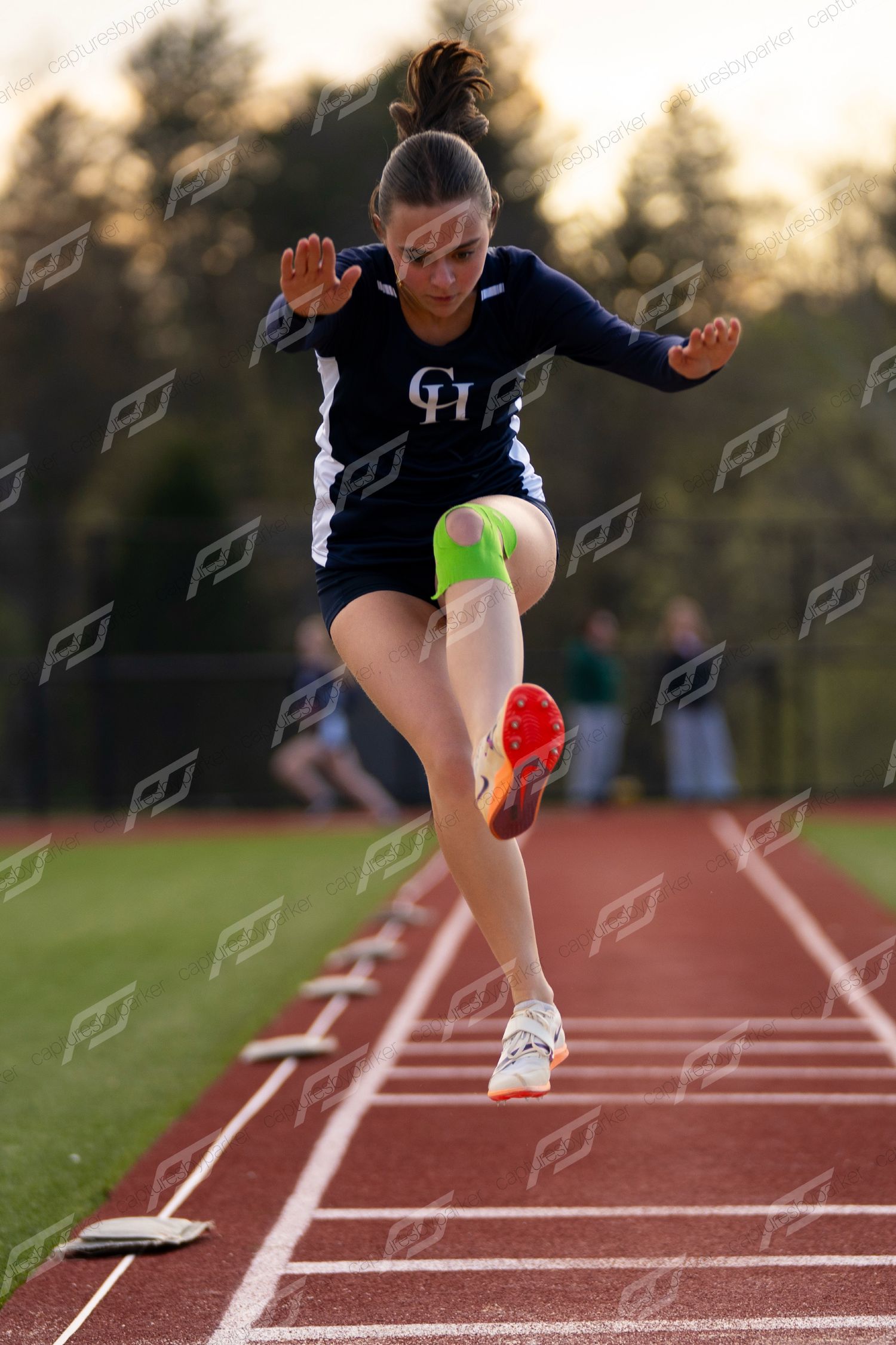 Female track athlete in navy CH uniform leaps powerfully down the runway during a jumping event.