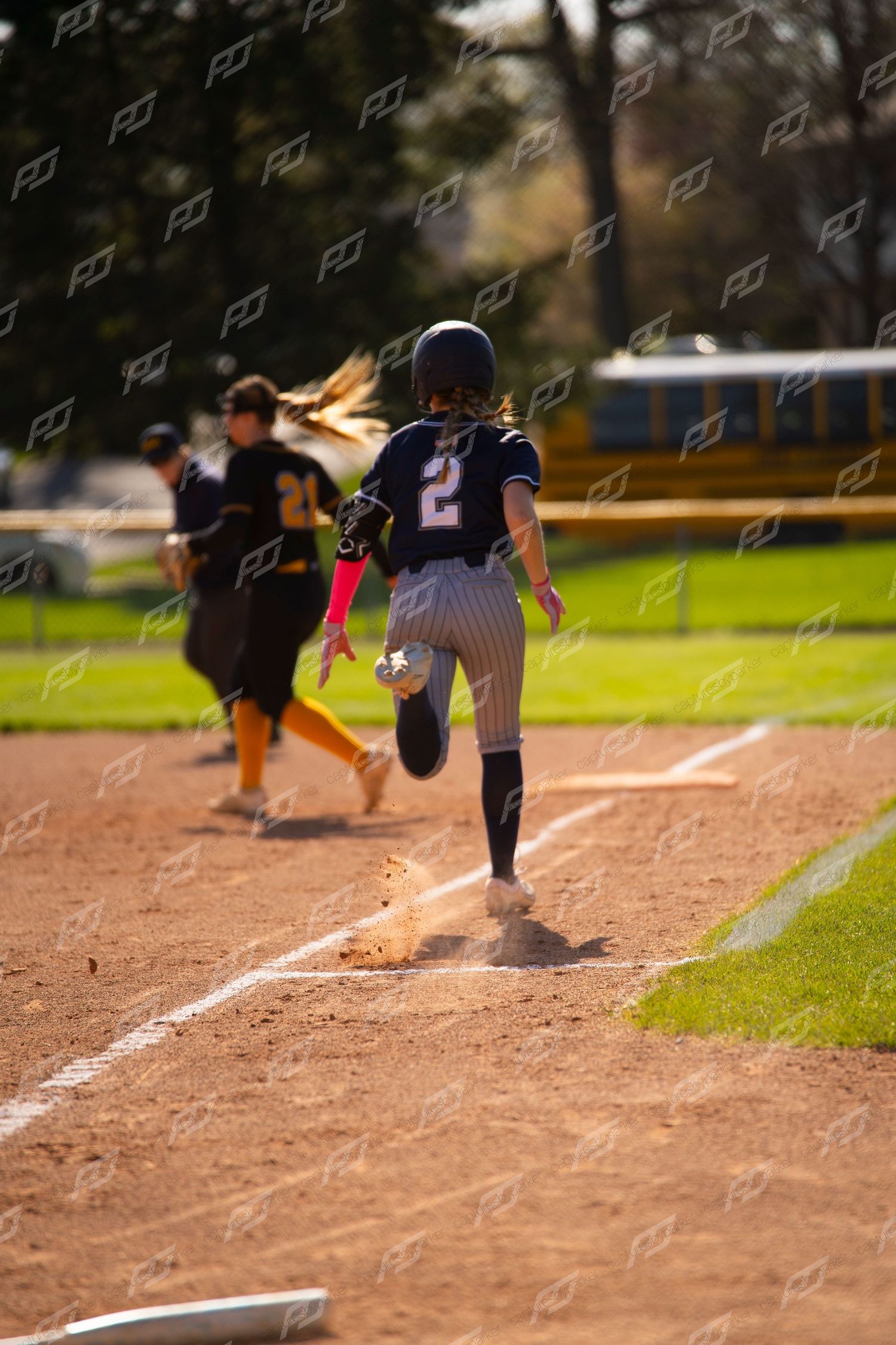 Softball player number 2 sprints down baseline as opposing player chases during sunny outdoor game.