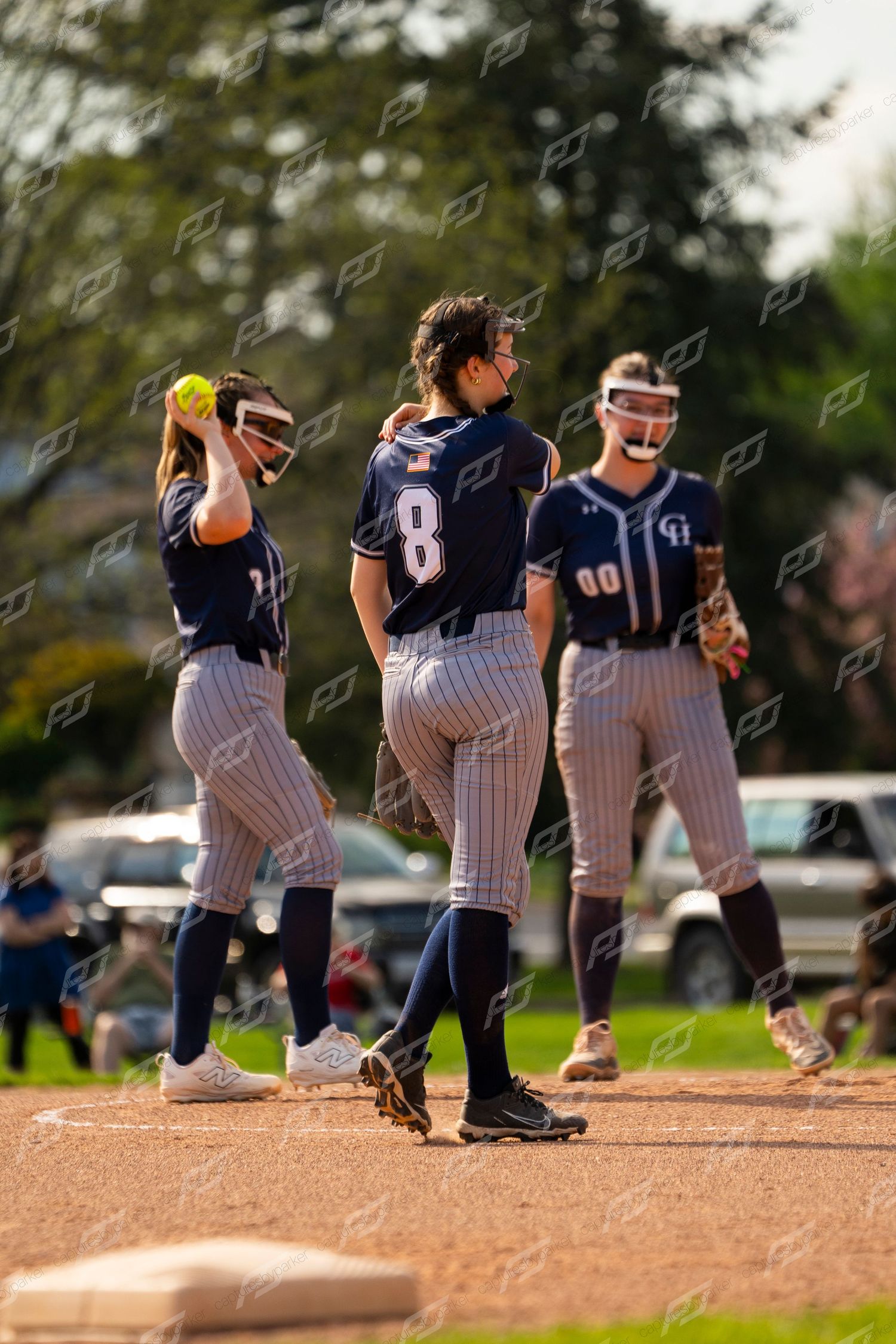 Three female softball players in navy pinstripe uniforms gather on field during pitching change.