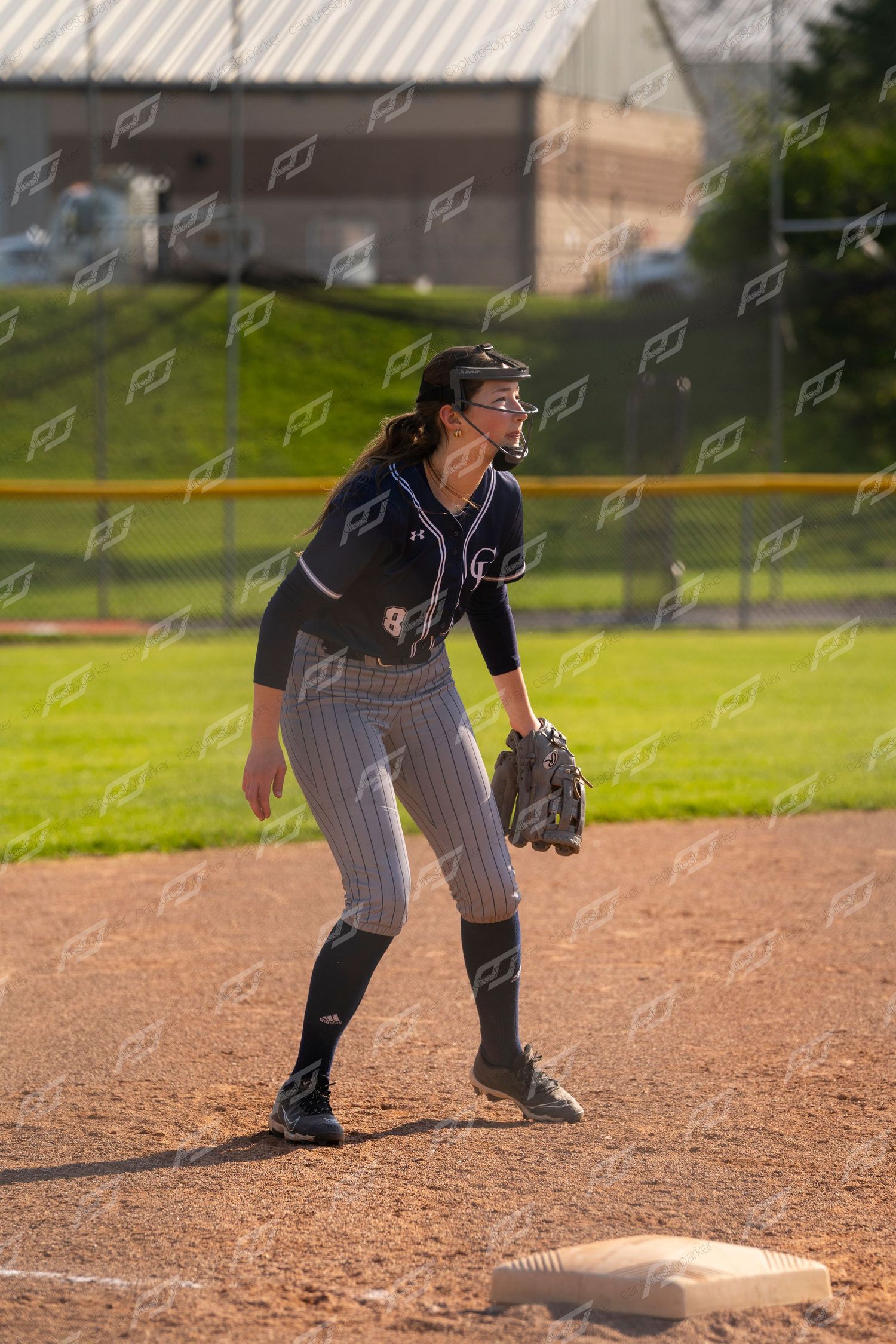 Female softball infielder in navy uniform crouching in ready position at first base during a game.
