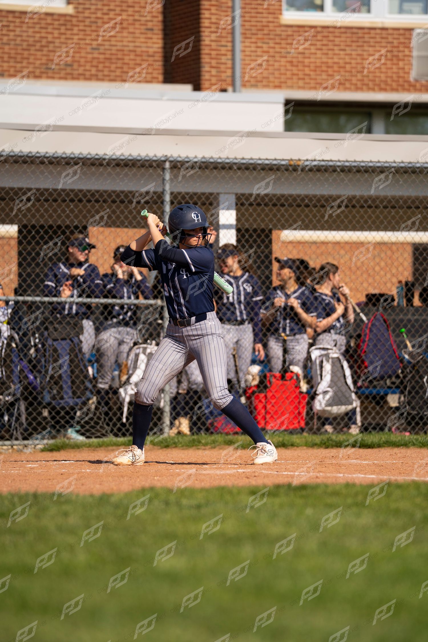 Softball player in navy pinstripe uniform winds up to bat at home plate with teammates visible in dugout behind chain-link fence.