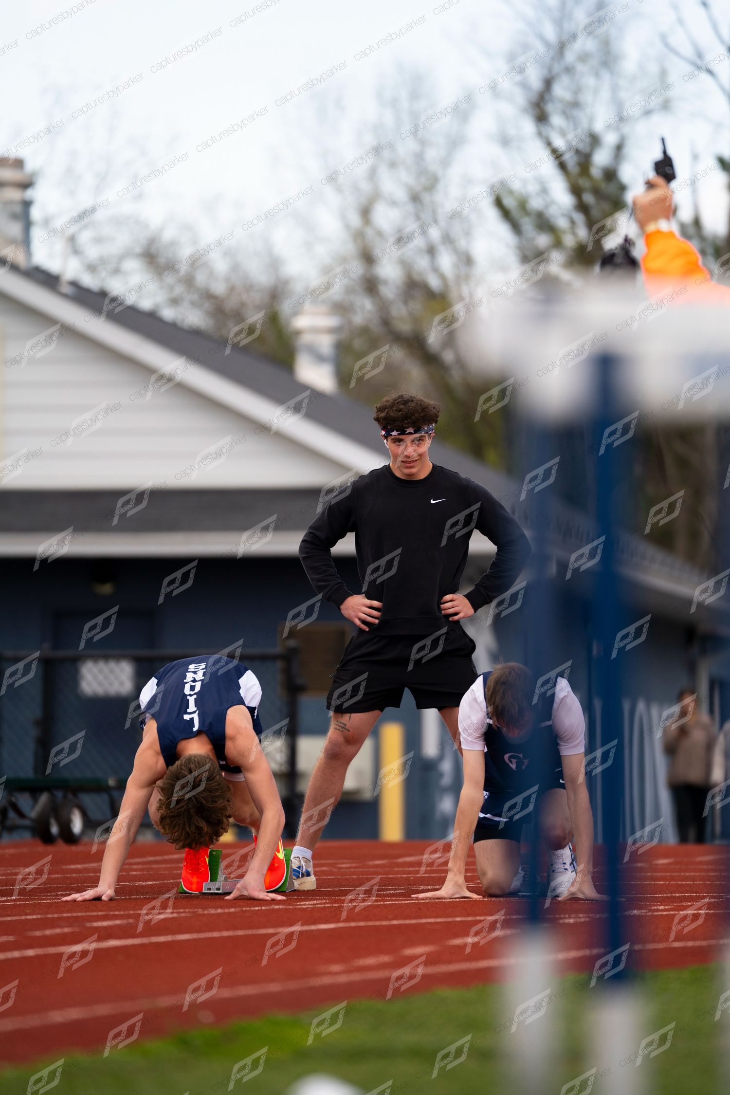 Male sprinter sets up in starting blocks as teammate watches at outdoor high school track meet.