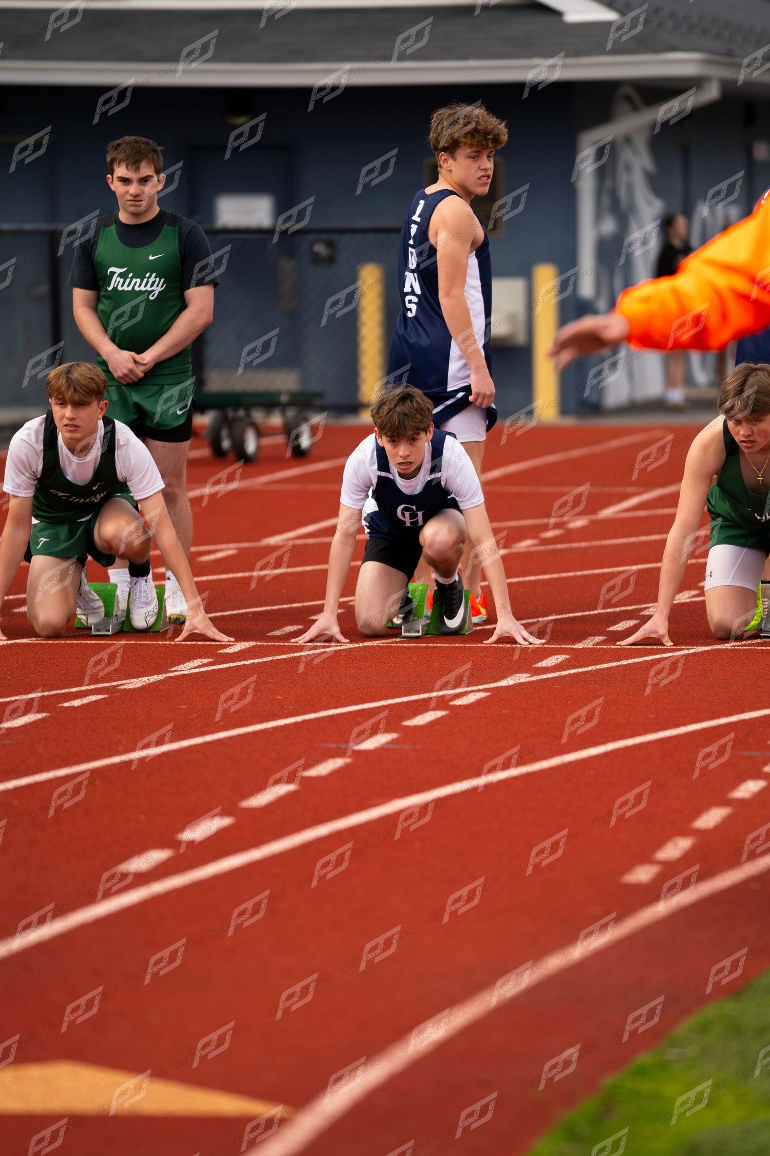High school sprinters set in starting blocks on red track with Lions and Trinity athletes competing.