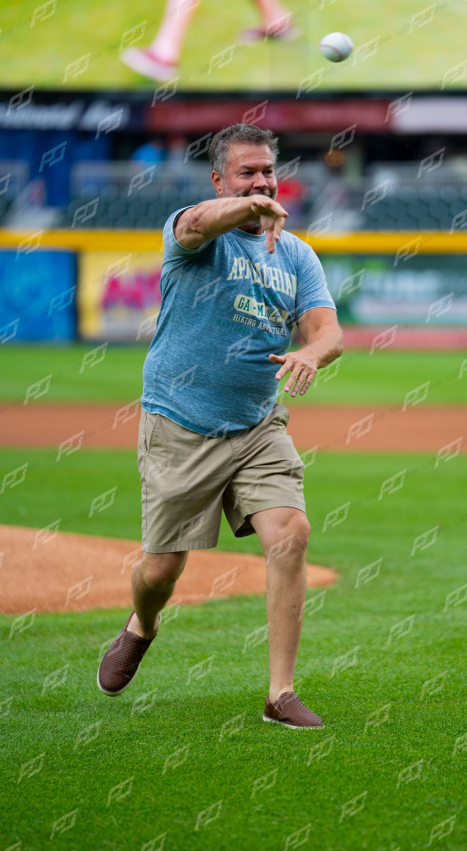 Man in casual clothes throwing a ceremonial first pitch on a baseball field at a minor league game.