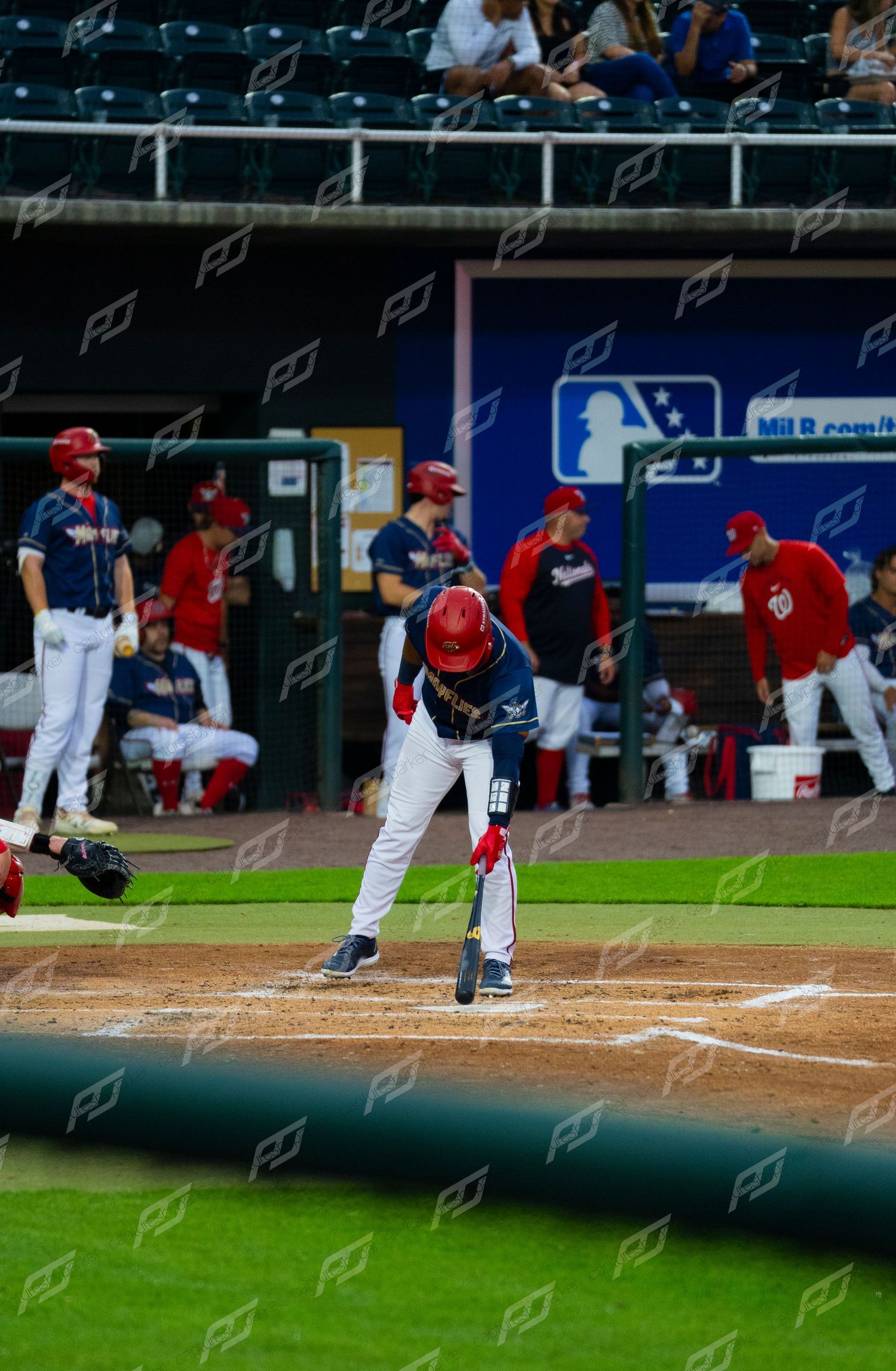 Baseball player in red helmet taps home plate with bat while teammates watch from dugout during game.