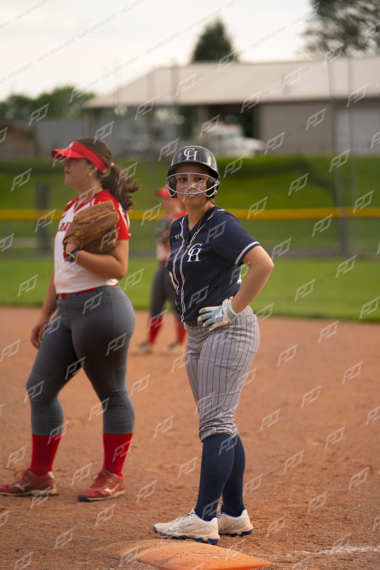 Female softball player standing on base in navy uniform beside red-uniformed fielder.
