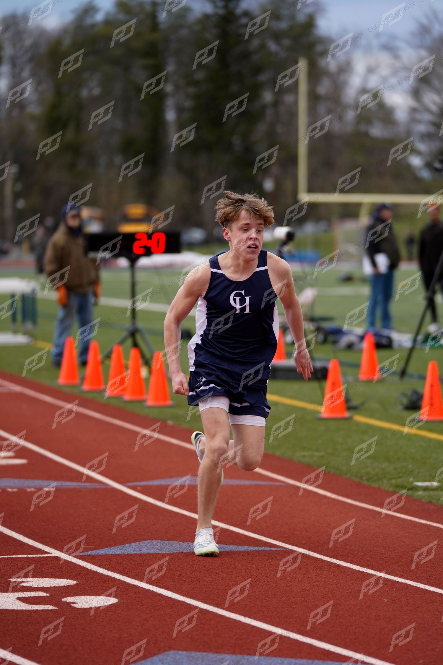 Male track athlete in navy CH uniform runs at full speed on red outdoor track during a meet.