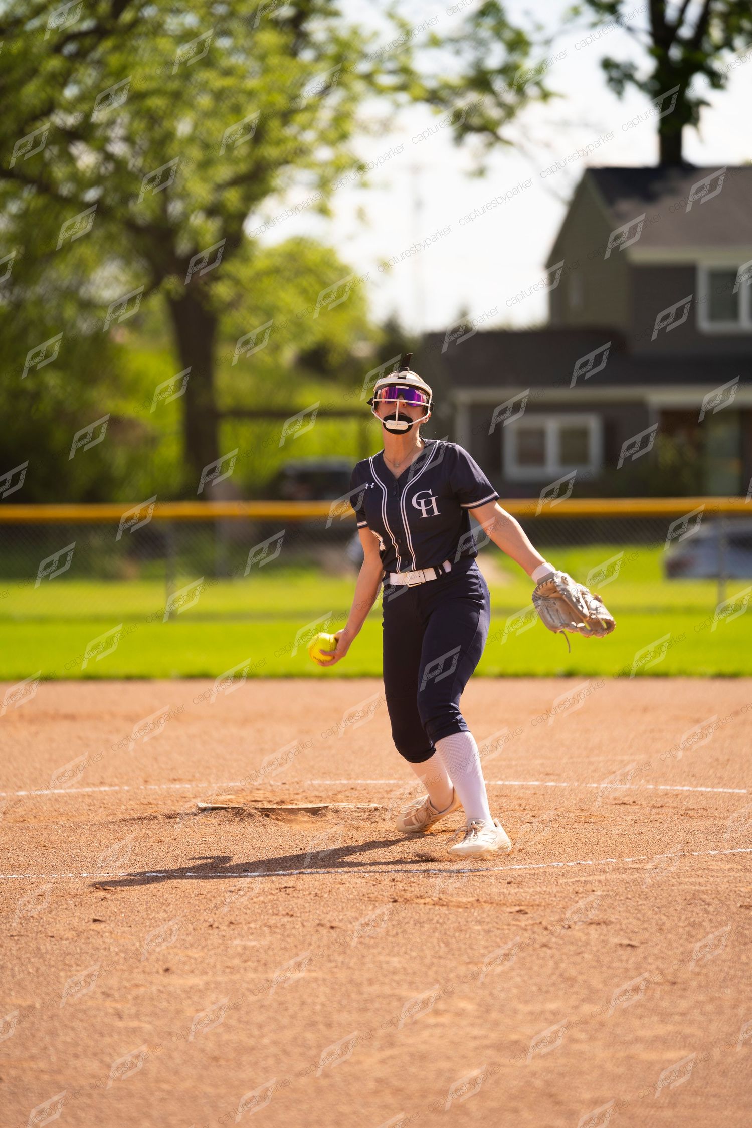 Female softball pitcher in navy uniform smiles while preparing to deliver pitch on mound.