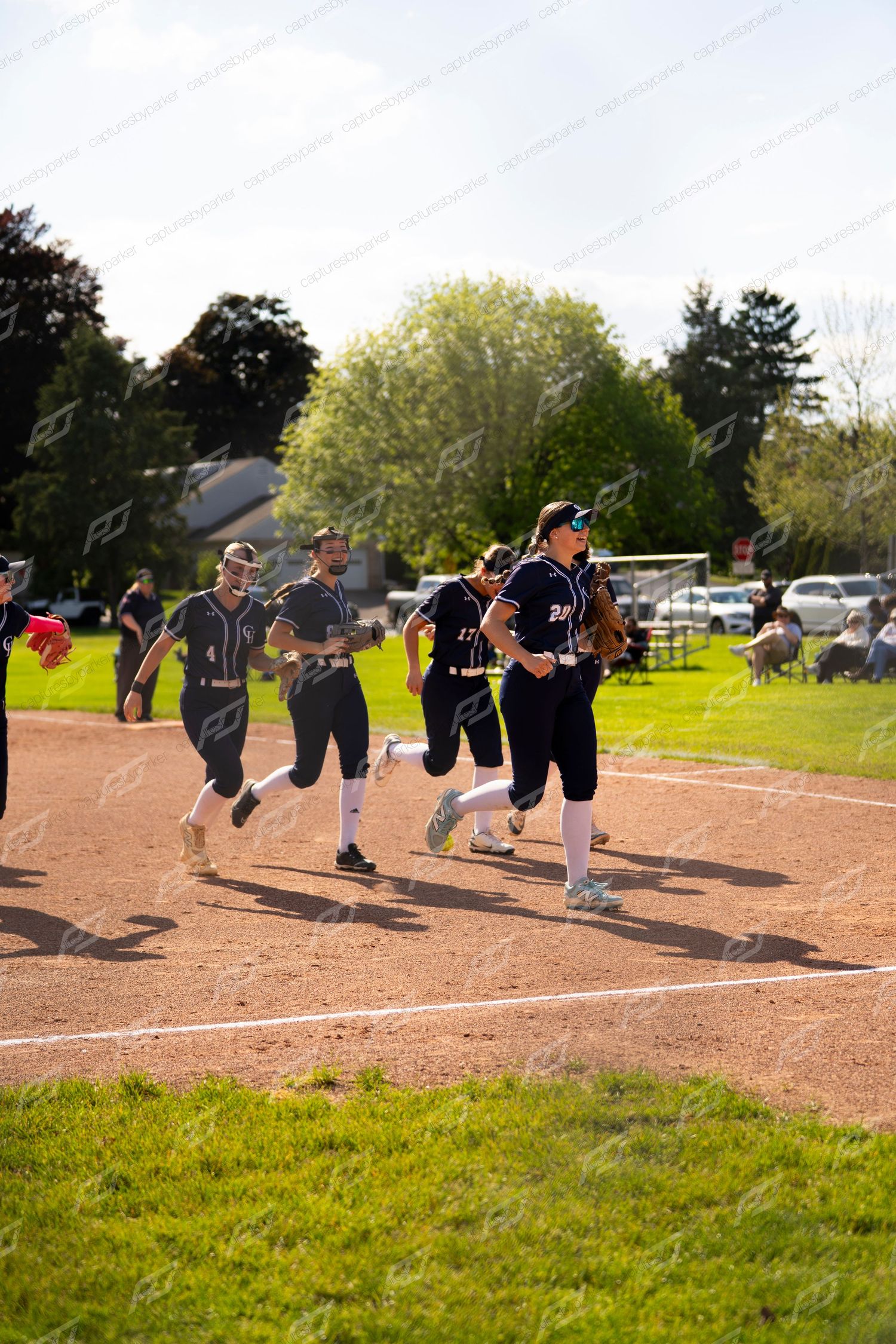 Softball players in dark uniforms running across the infield dirt during a sunny outdoor game.