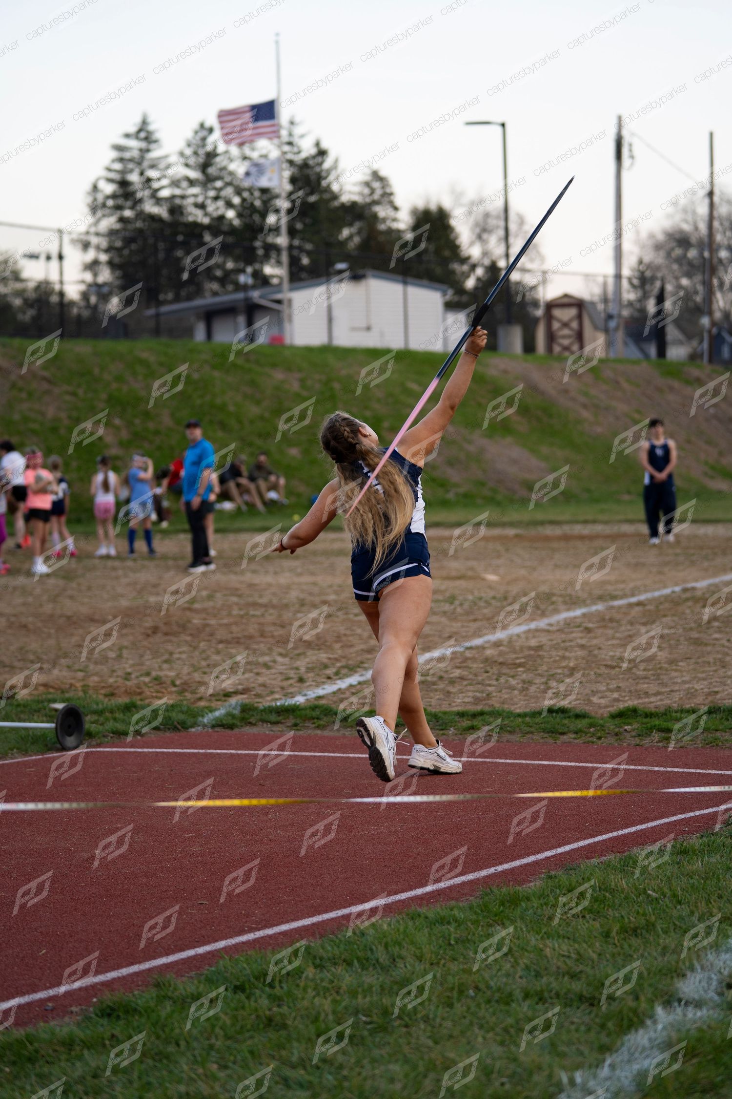 Female athlete in navy uniform throwing javelin at outdoor track meet.