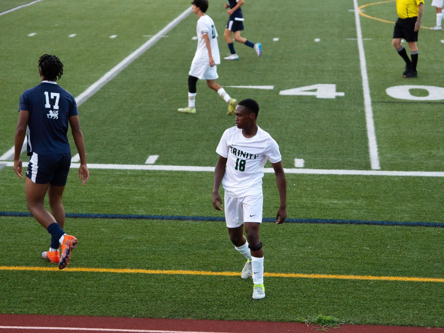 Trinity soccer player number 18 in white uniform runs on turf field during night game.