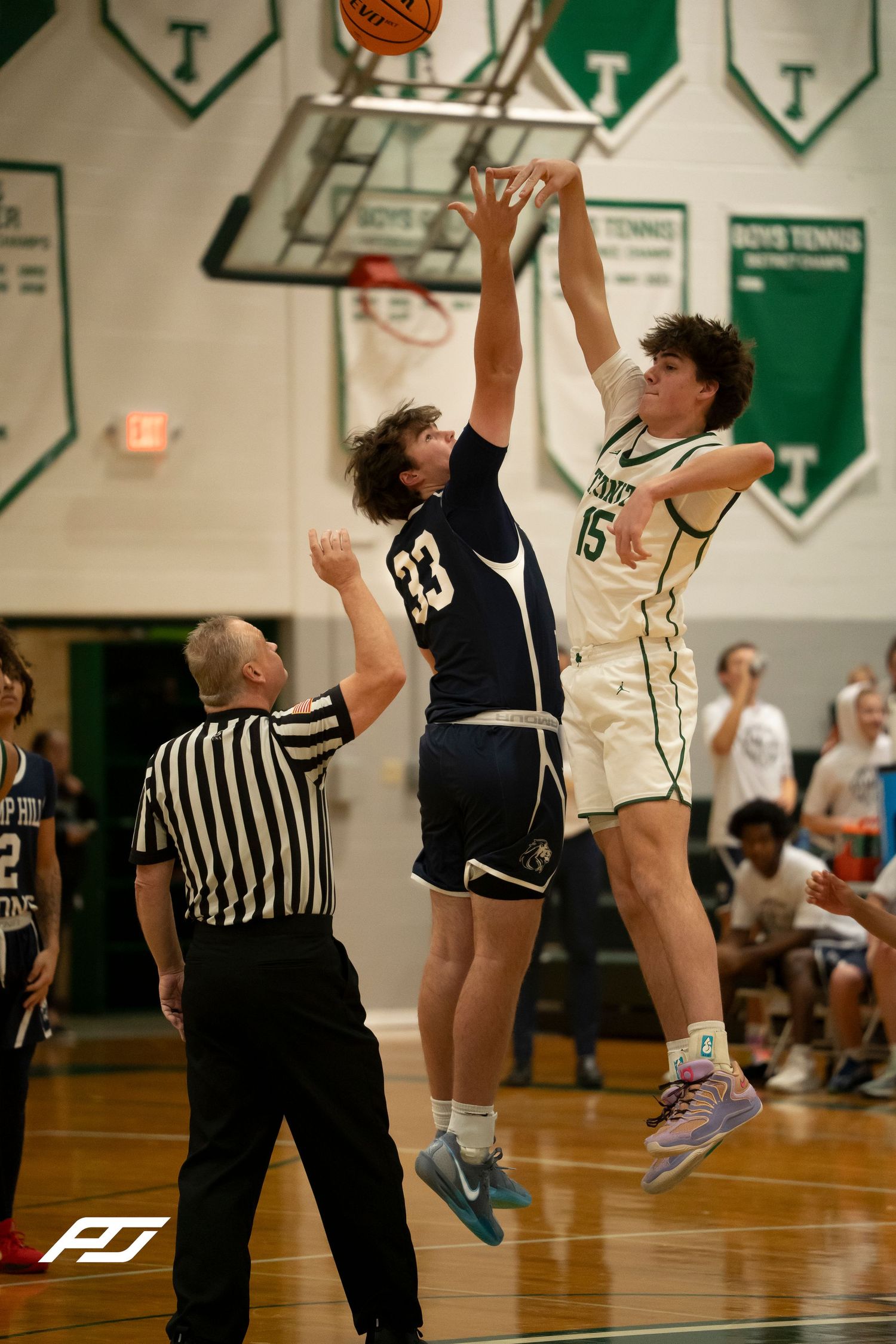 Two players jump for tip-off as referee watches during a high school basketball game in green gymnasium.