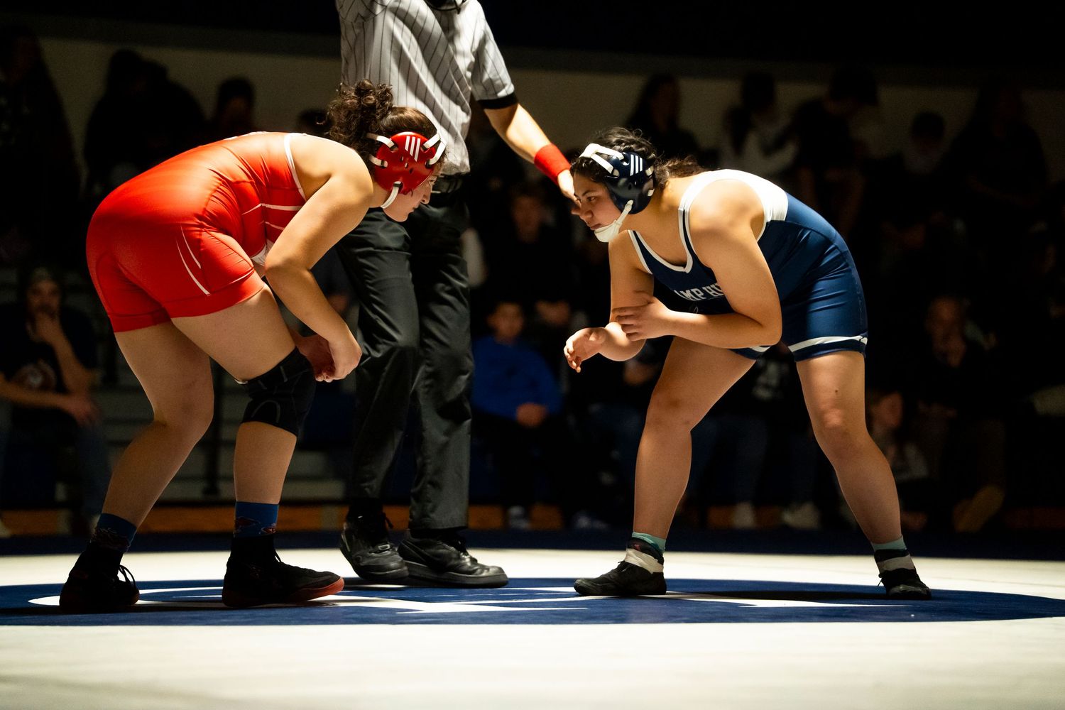 Two heavyweight wrestlers in red and blue singlets face off before referee at start of wrestling match.