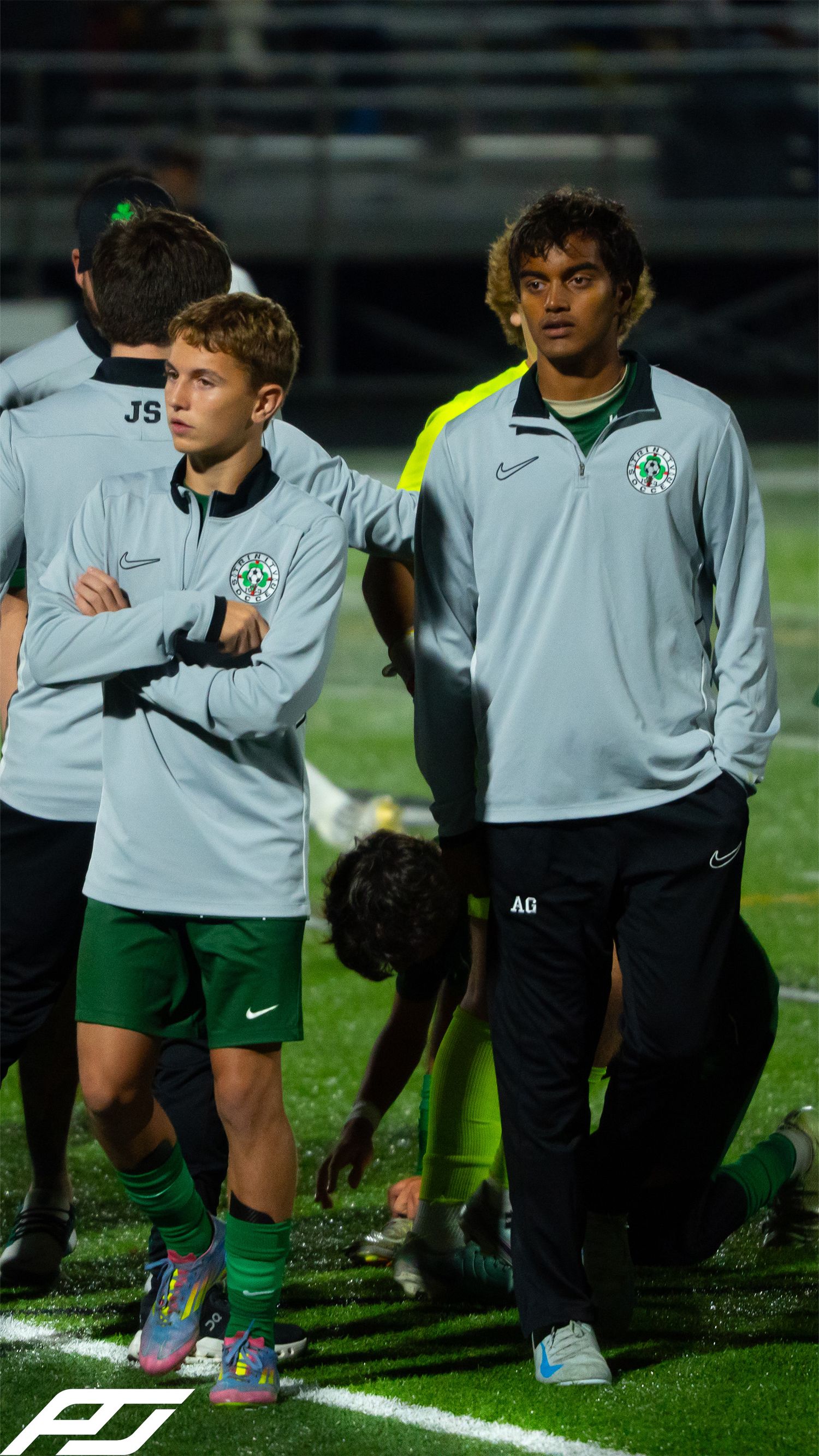 Two Trinity soccer team staff members in gray Nike jackets stand on the sideline during a night match.