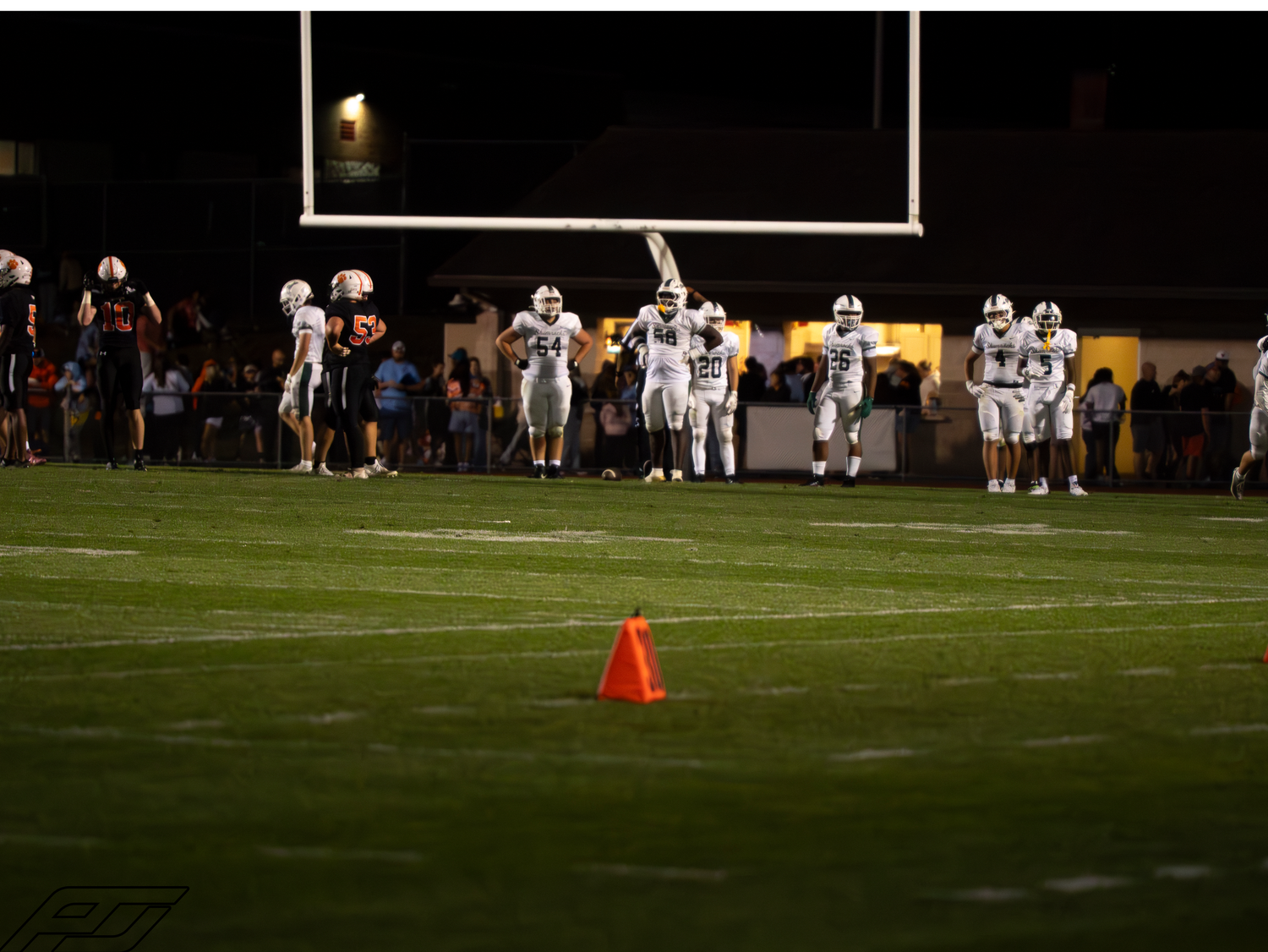 High school football players in white uniforms stand on a green field at night with a orange yard marker cone visible.