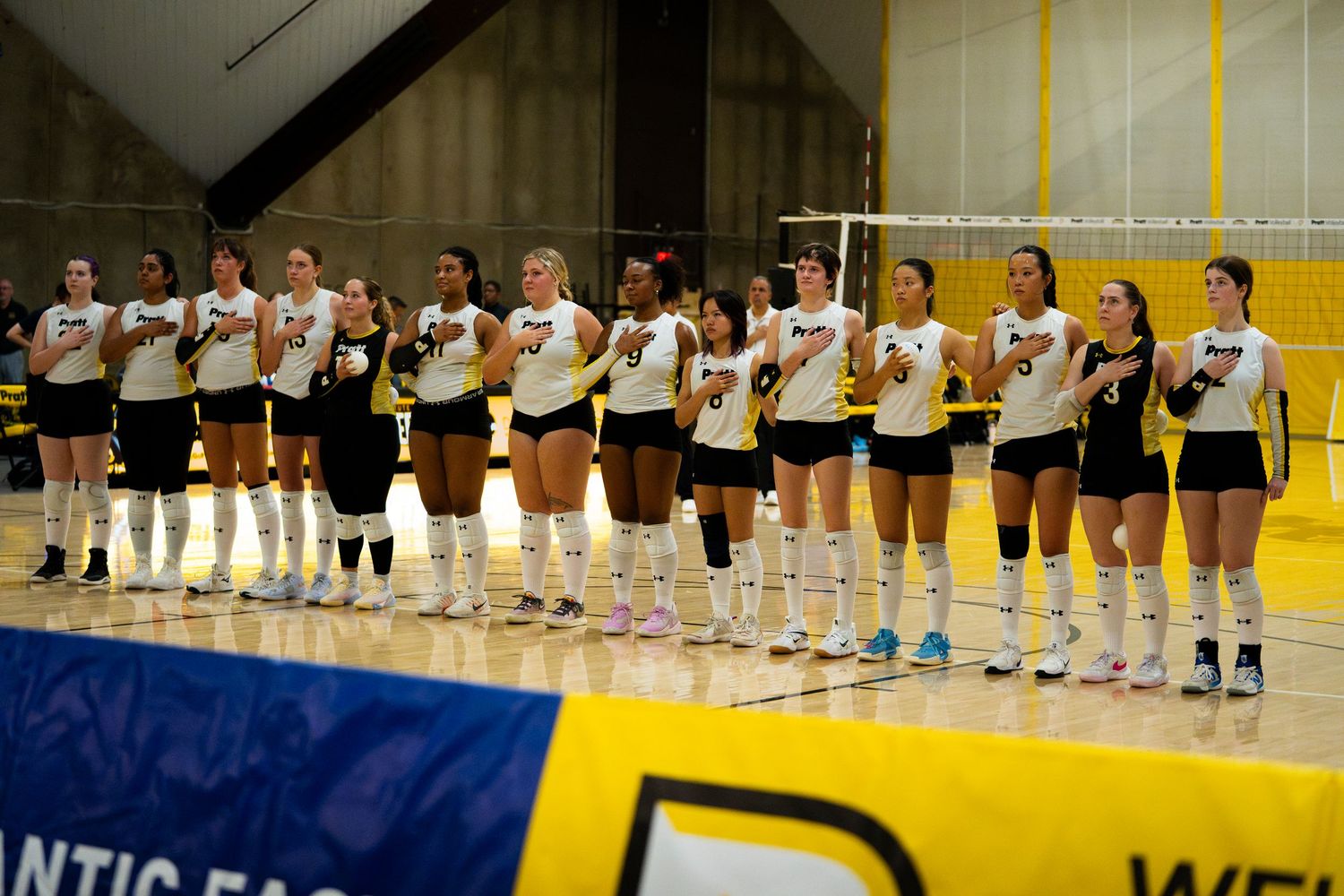 Women's volleyball team lined up on indoor court in white uniforms during pre-match ceremony at Atlantic East conference game.