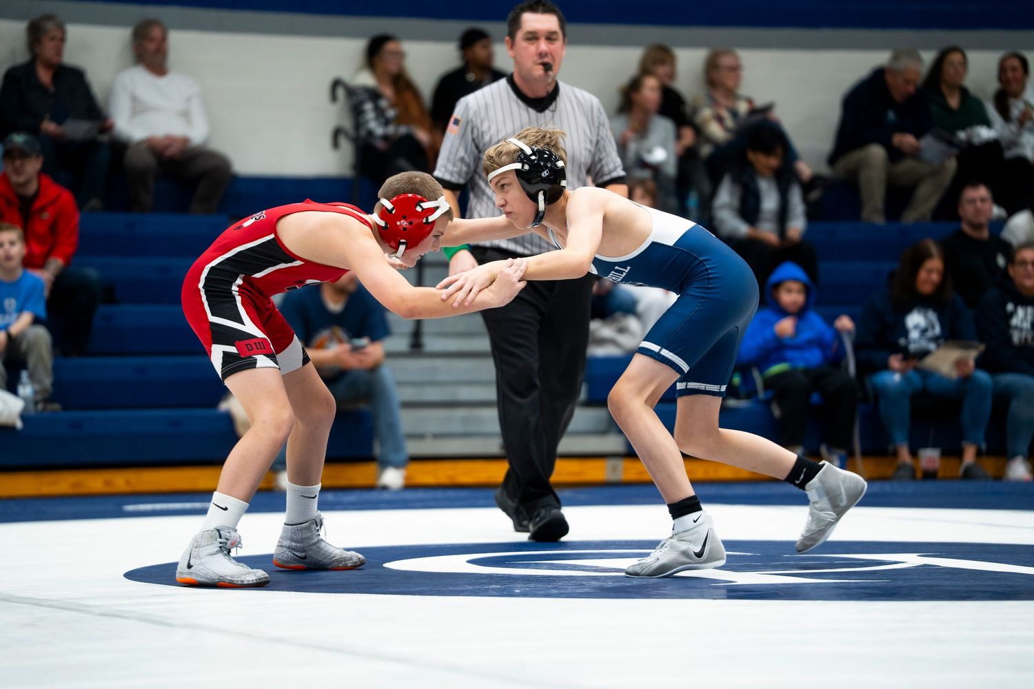 Two wrestlers in red and blue singlets face off on a wrestling mat during a high school match.