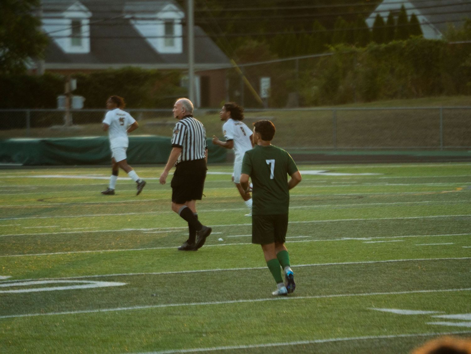 Soccer player number 7 in green uniform runs alongside a referee and opponent on a turf field.