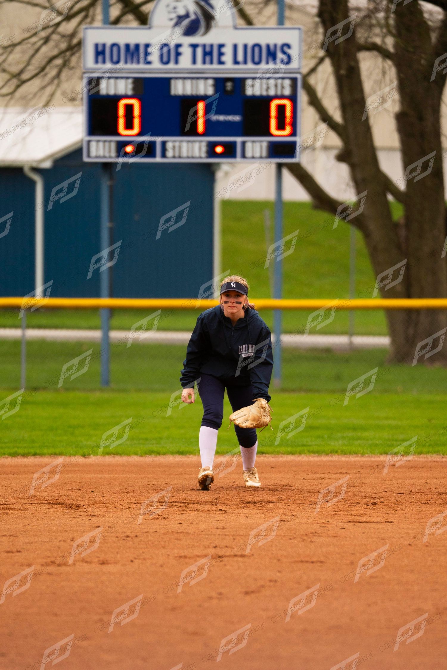 Softball infielder in navy uniform fielding a ground ball on a red clay diamond with Lions scoreboard in background.