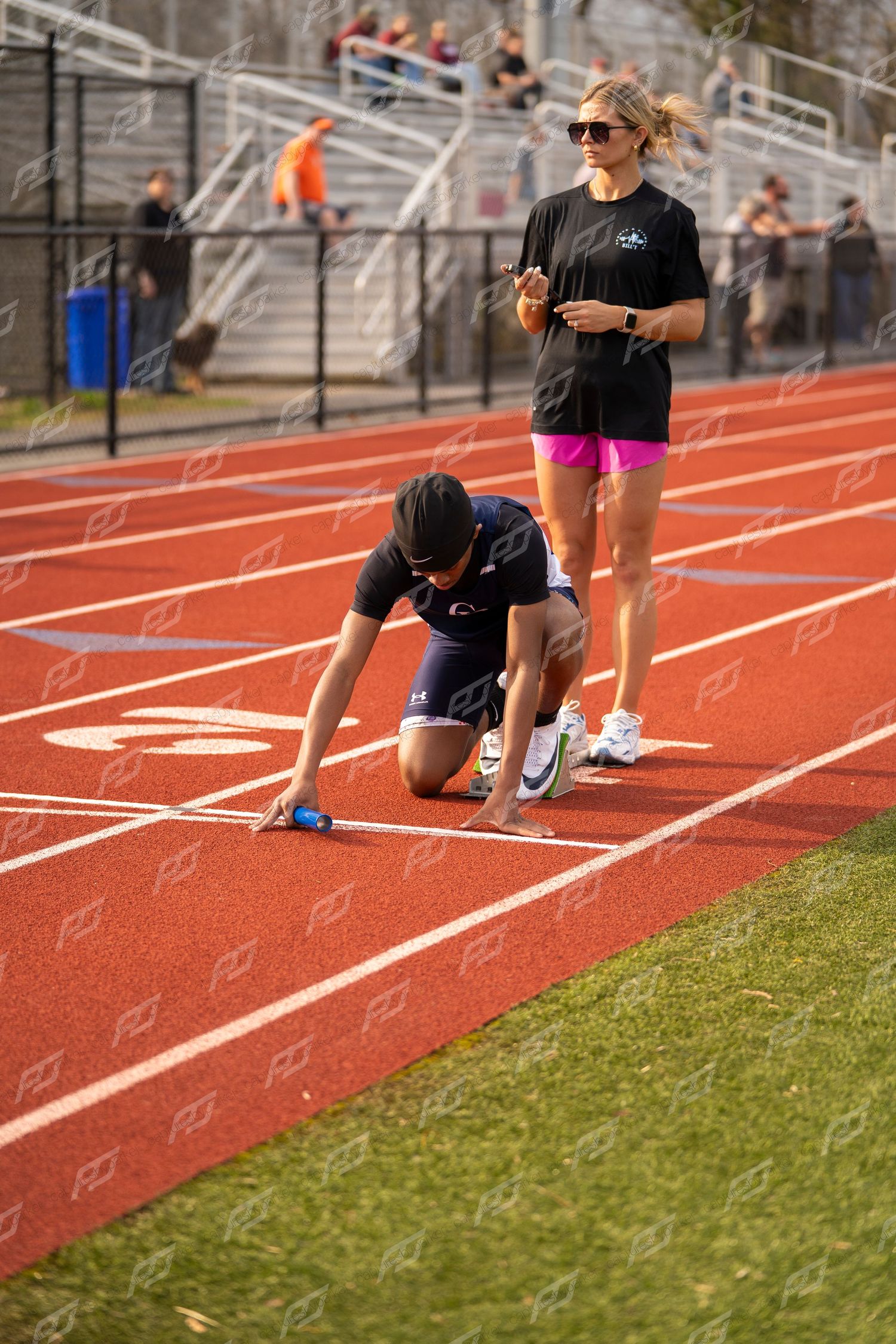 Sprinter sets starting blocks on red track lane as female coach stands nearby watching.