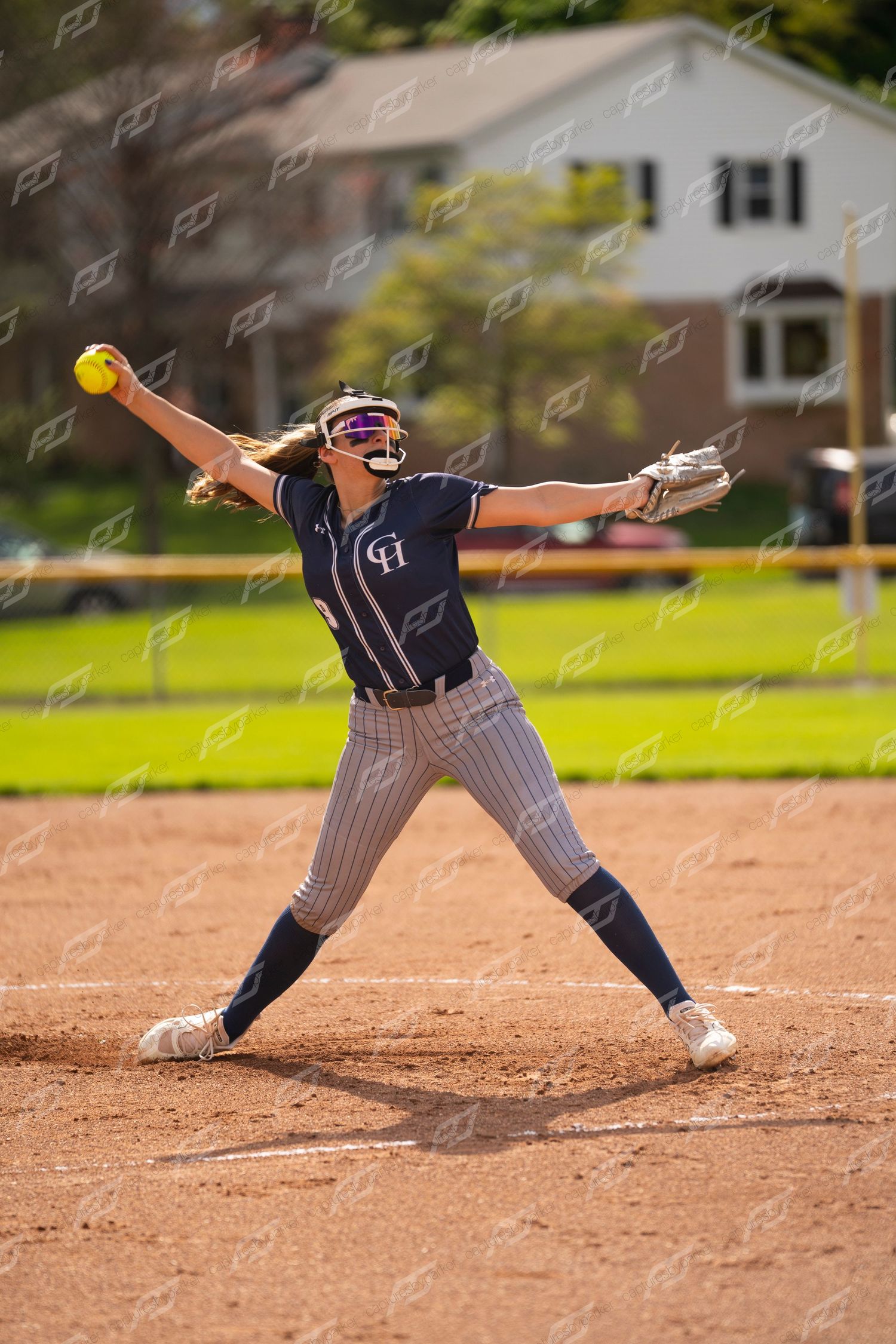 Female softball pitcher in navy uniform winds up to throw yellow ball during outdoor game.