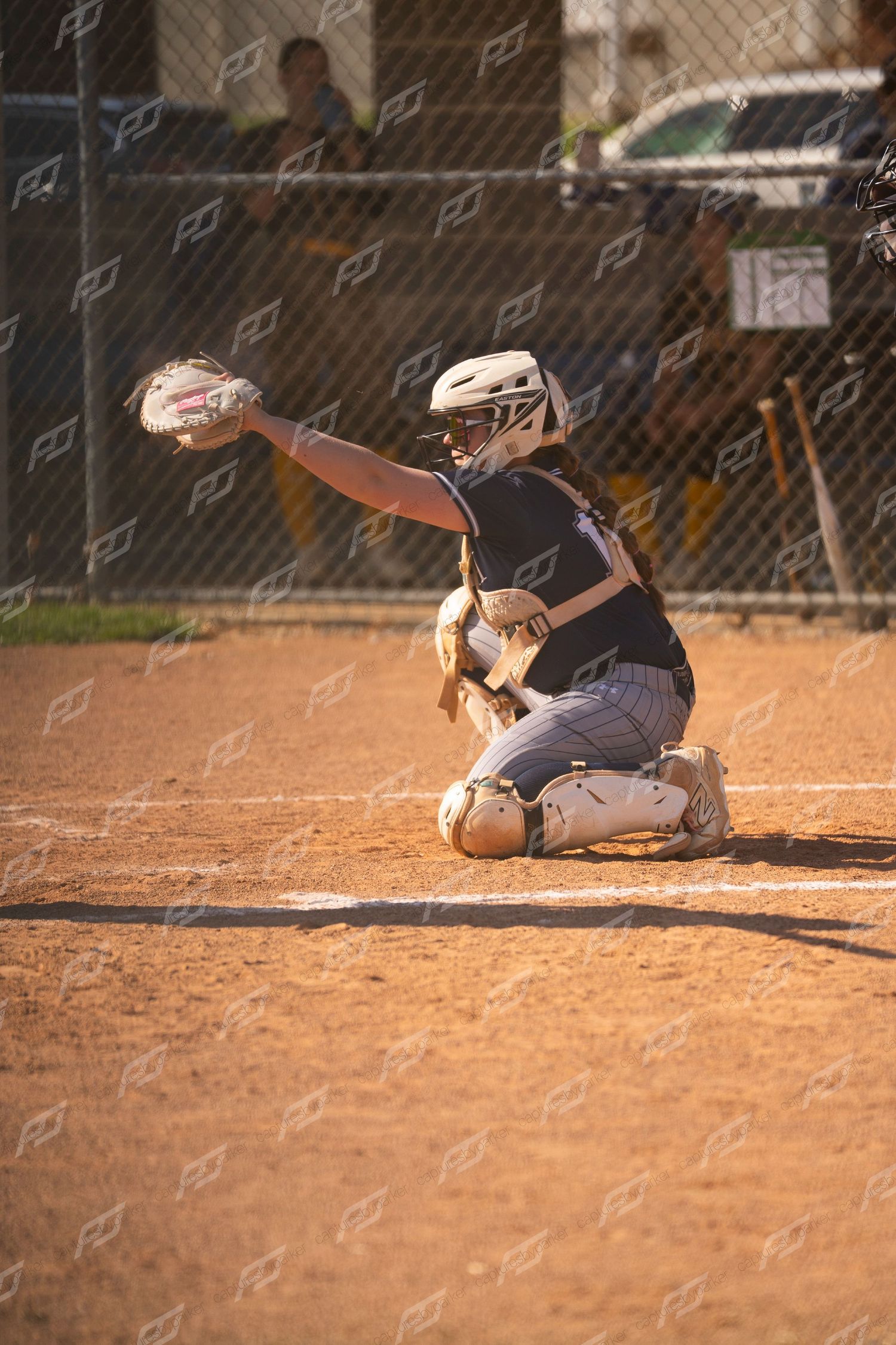 Softball catcher in dark uniform kneels in dirt and reaches glove out to receive a pitch.