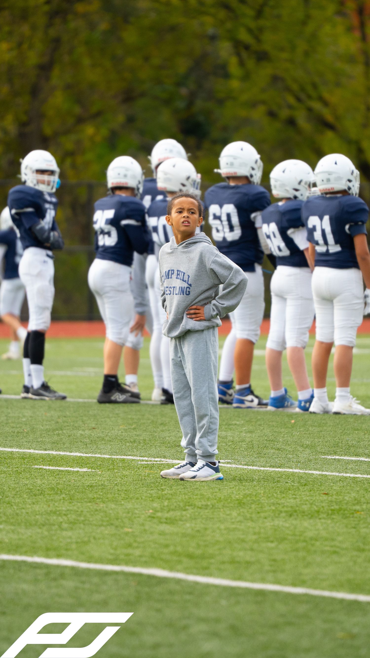 Young boy in Camp Hill wrestling hoodie stands before navy football team.