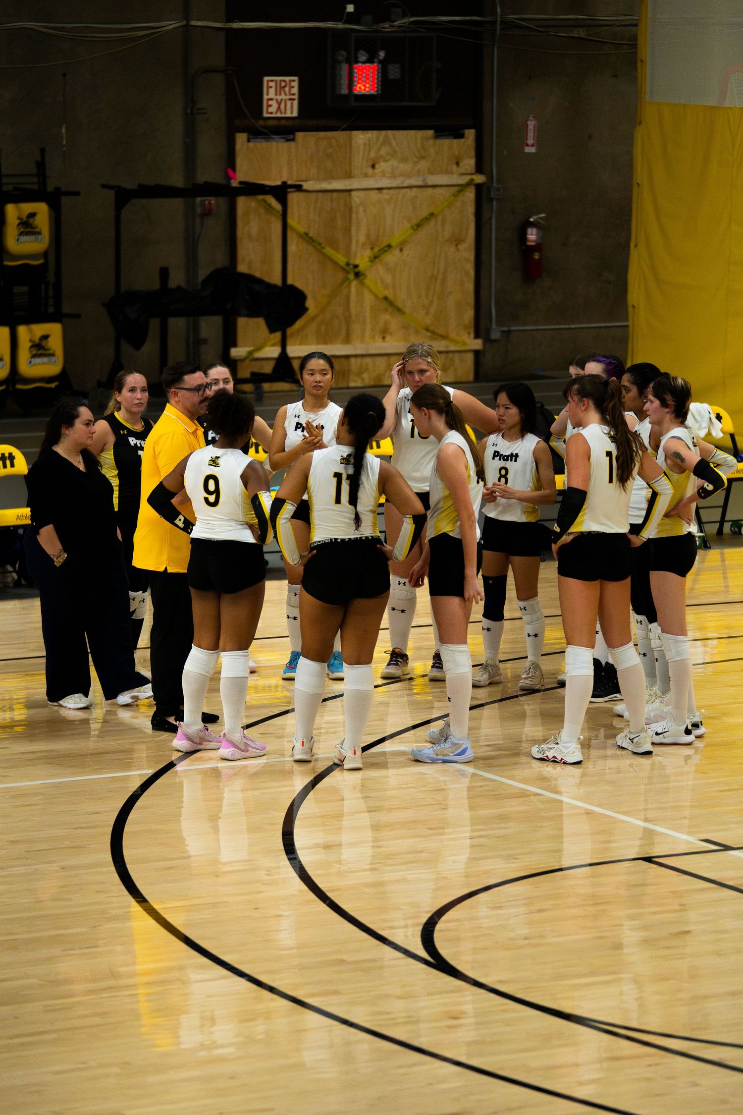 Volleyball team huddles on court in black and white uniforms, with coaches during a timeout in an indoor gymnasium.