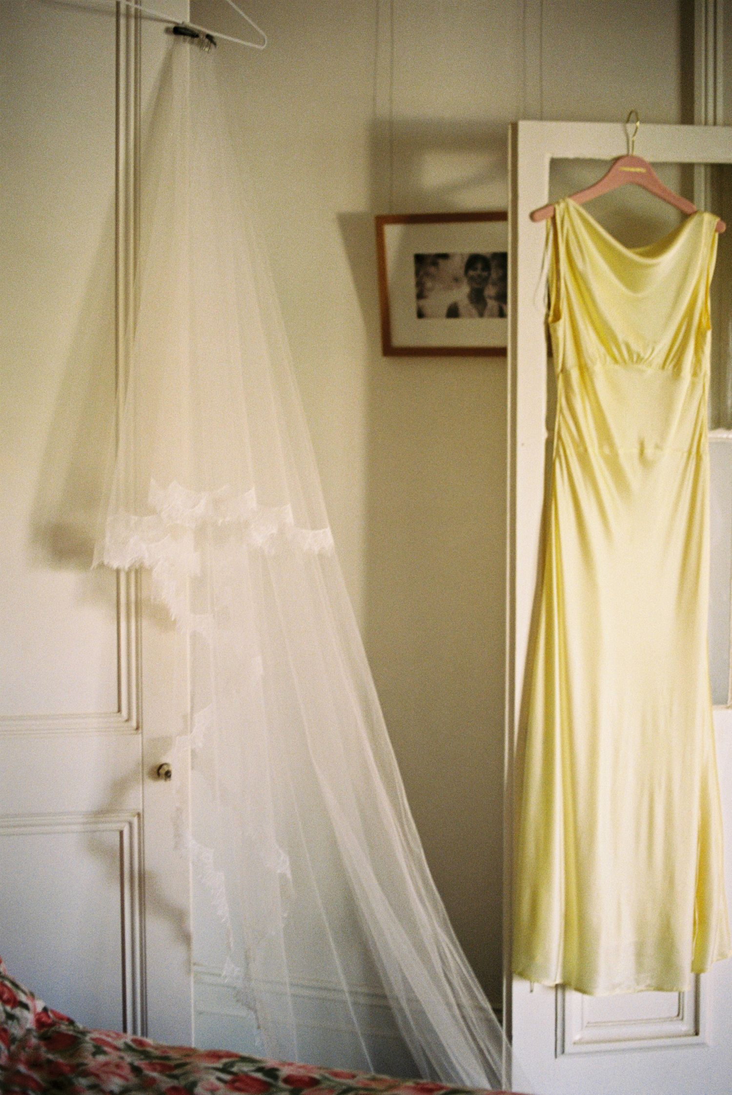 A pale yellow dress and white veil hang against a neutral wall in preparation for a special occasion.