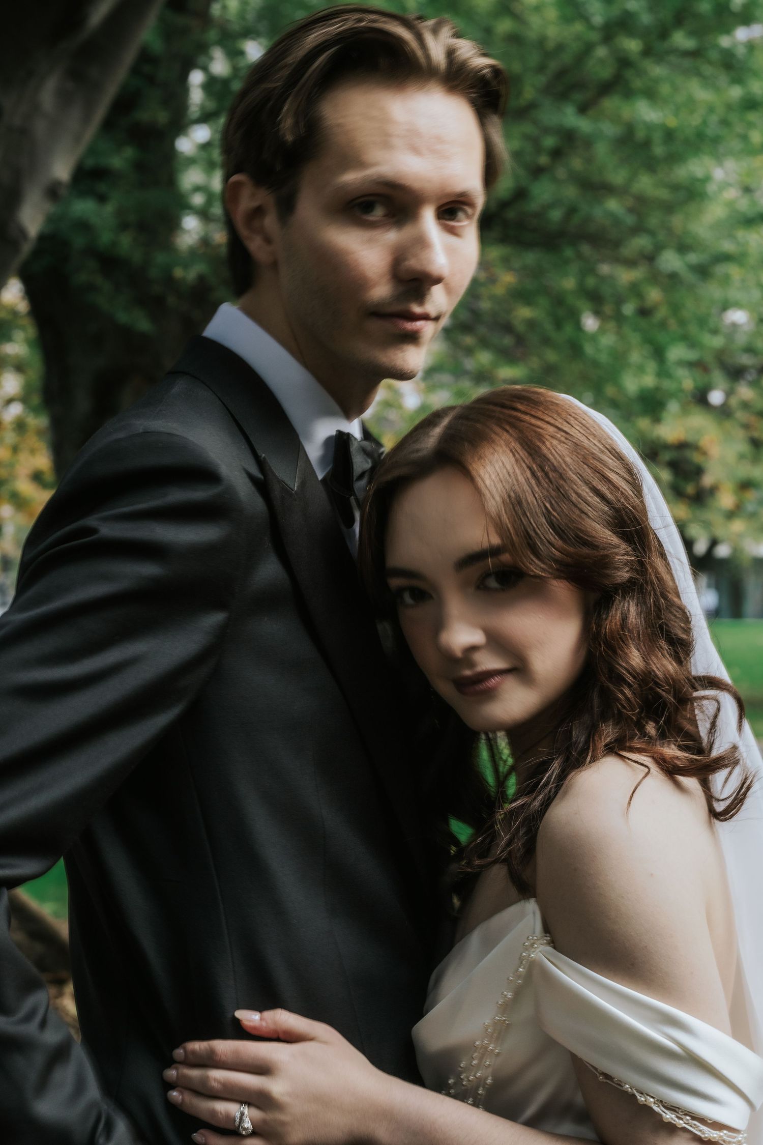 A bride and groom pose outdoors in a lush green park, wearing wedding attire and embracing for a romantic portrait.