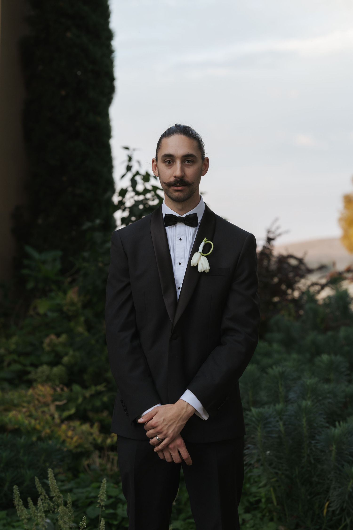 Groom in black tuxedo with white boutonniere stands outdoors among lush greenery at dusk.