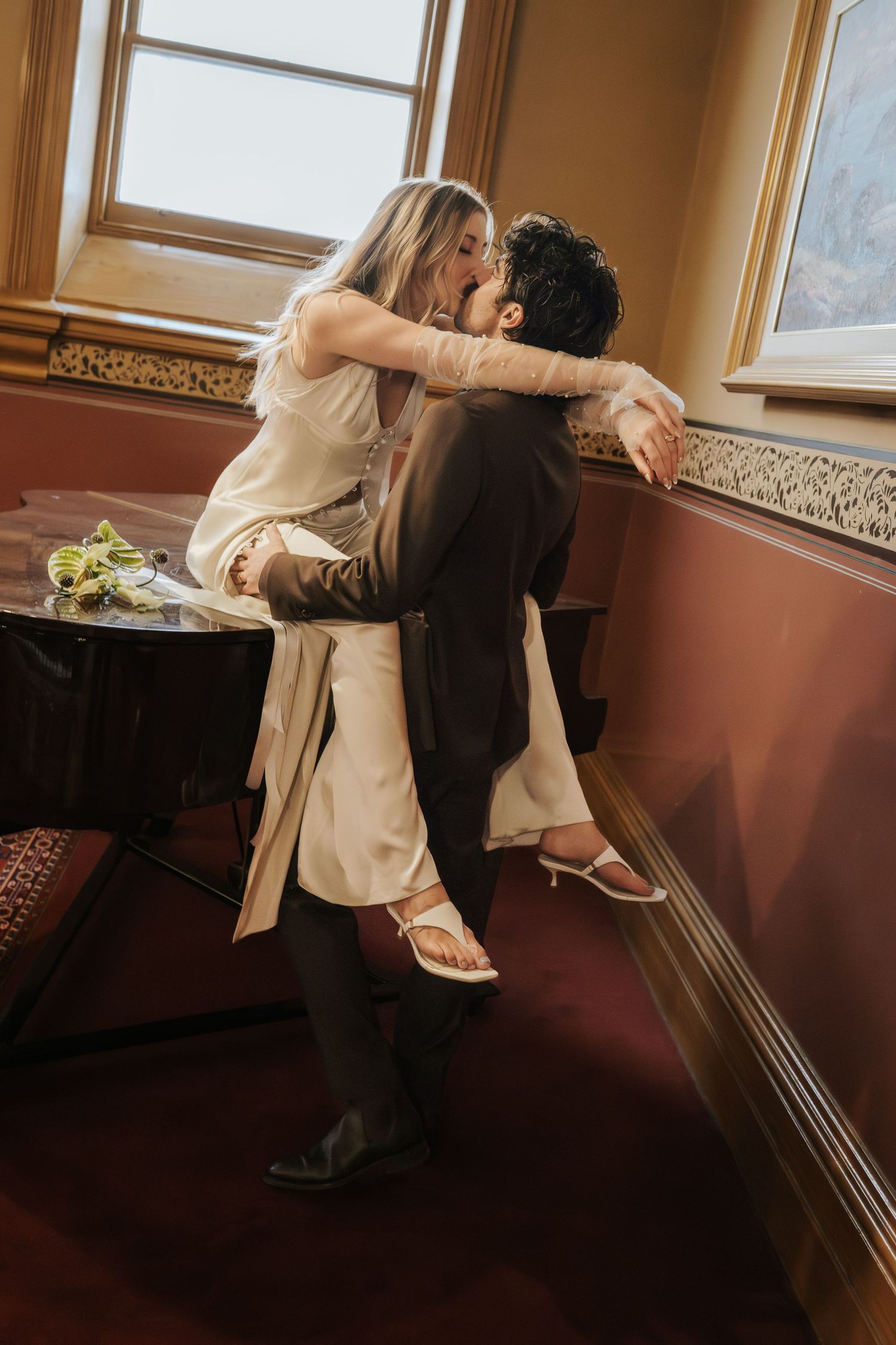 A couple in wedding attire shares a romantic kiss, with the groom lifting the bride near a window.