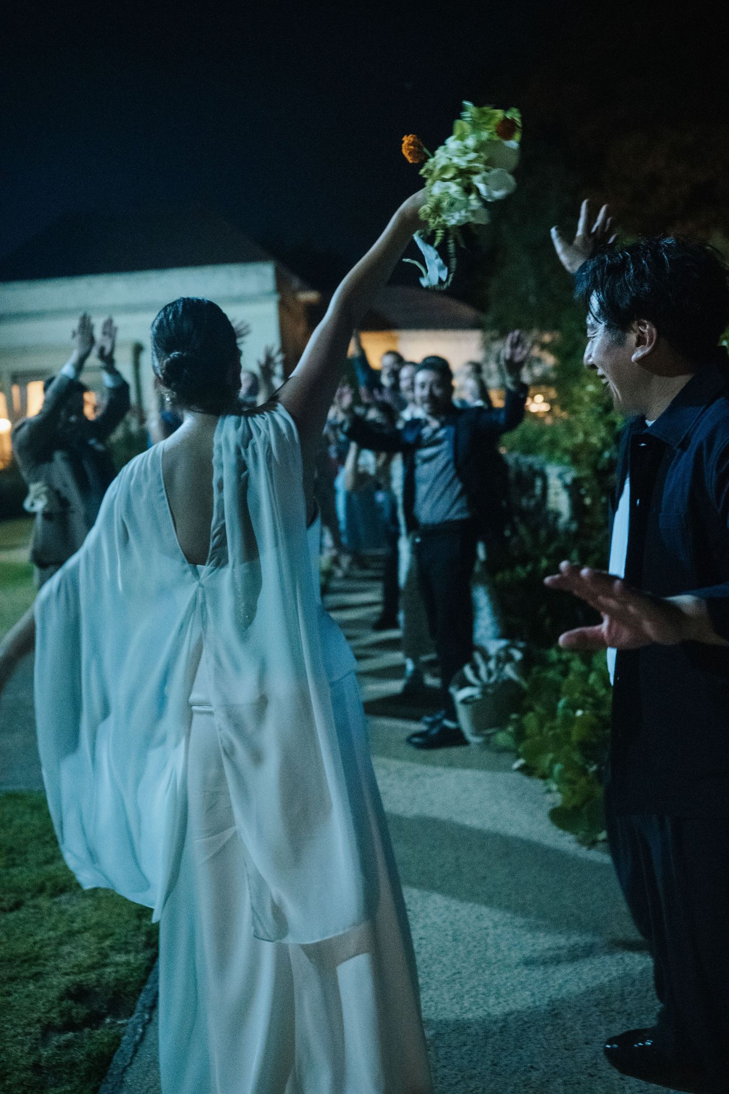 Bride in white gown raises bouquet to cheering wedding guests outdoors at night.