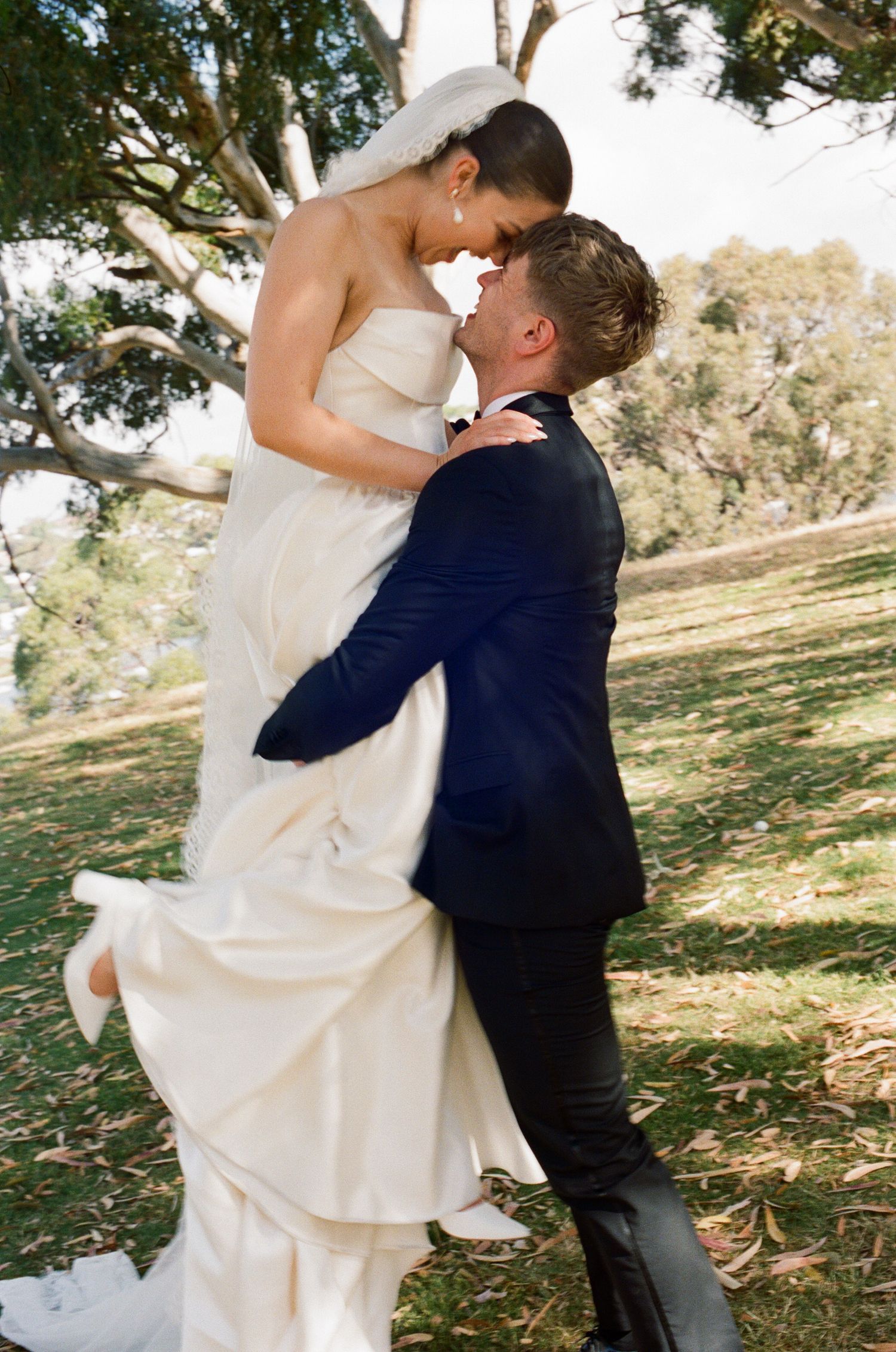 Couple in wedding attire embraces outdoors among trees.