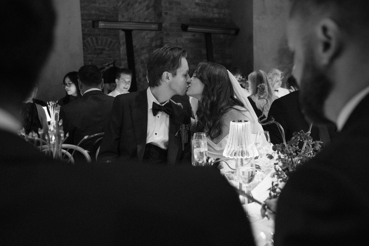 Black and white photo of a bride and groom sharing a kiss at their wedding reception dinner.