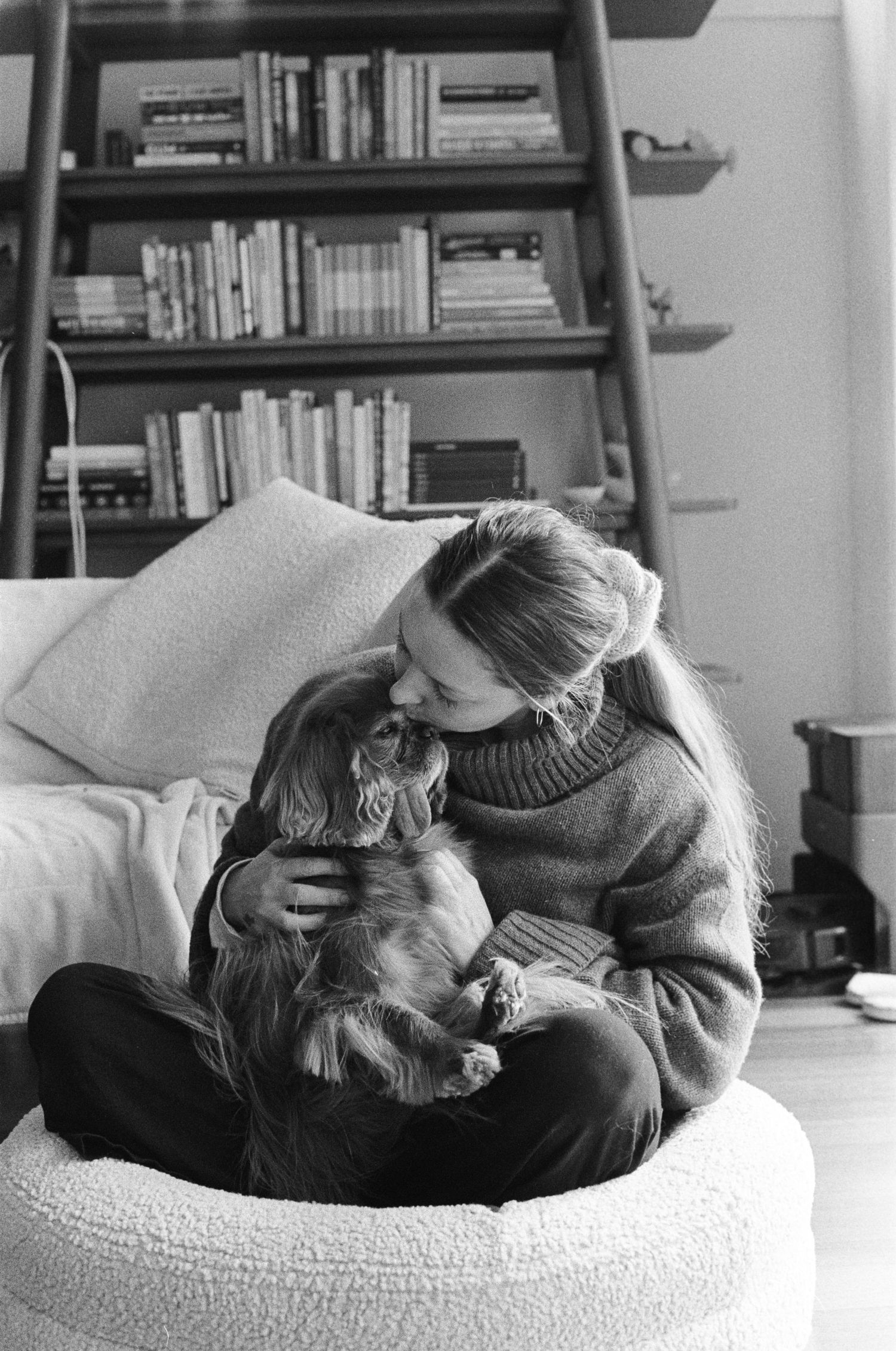 A cozy black and white scene shows someone in a turtleneck sweater cuddling with a dog in a plush chair near a bookshelf.