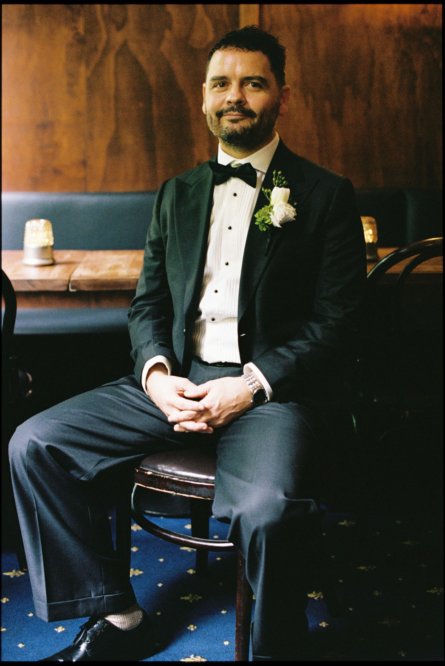 A groom in a black tuxedo with bow tie and boutonniere sits confidently in a classic wooden chair.