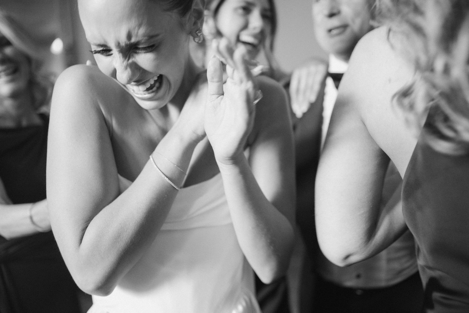 Black and white photo of a bride laughing and clapping joyfully with guests celebrating at a wedding reception.