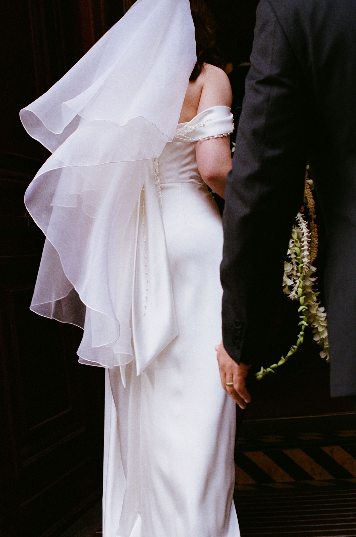 Bride in elegant white satin off-shoulder gown with flowing veil holding floral bouquet beside groom in gray suit.