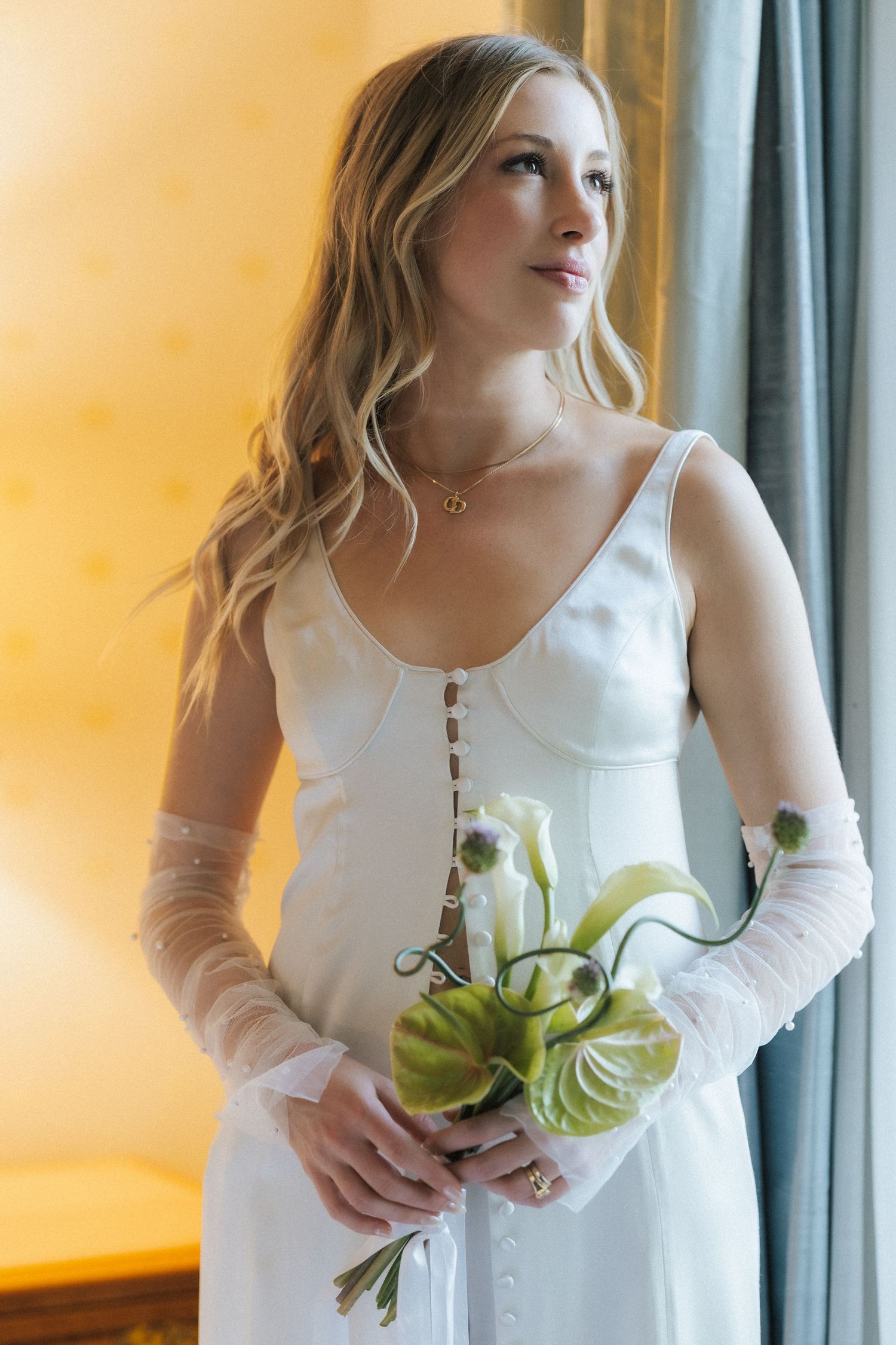 Bride in white satin gown with sheer gloves holds green bouquet by window.