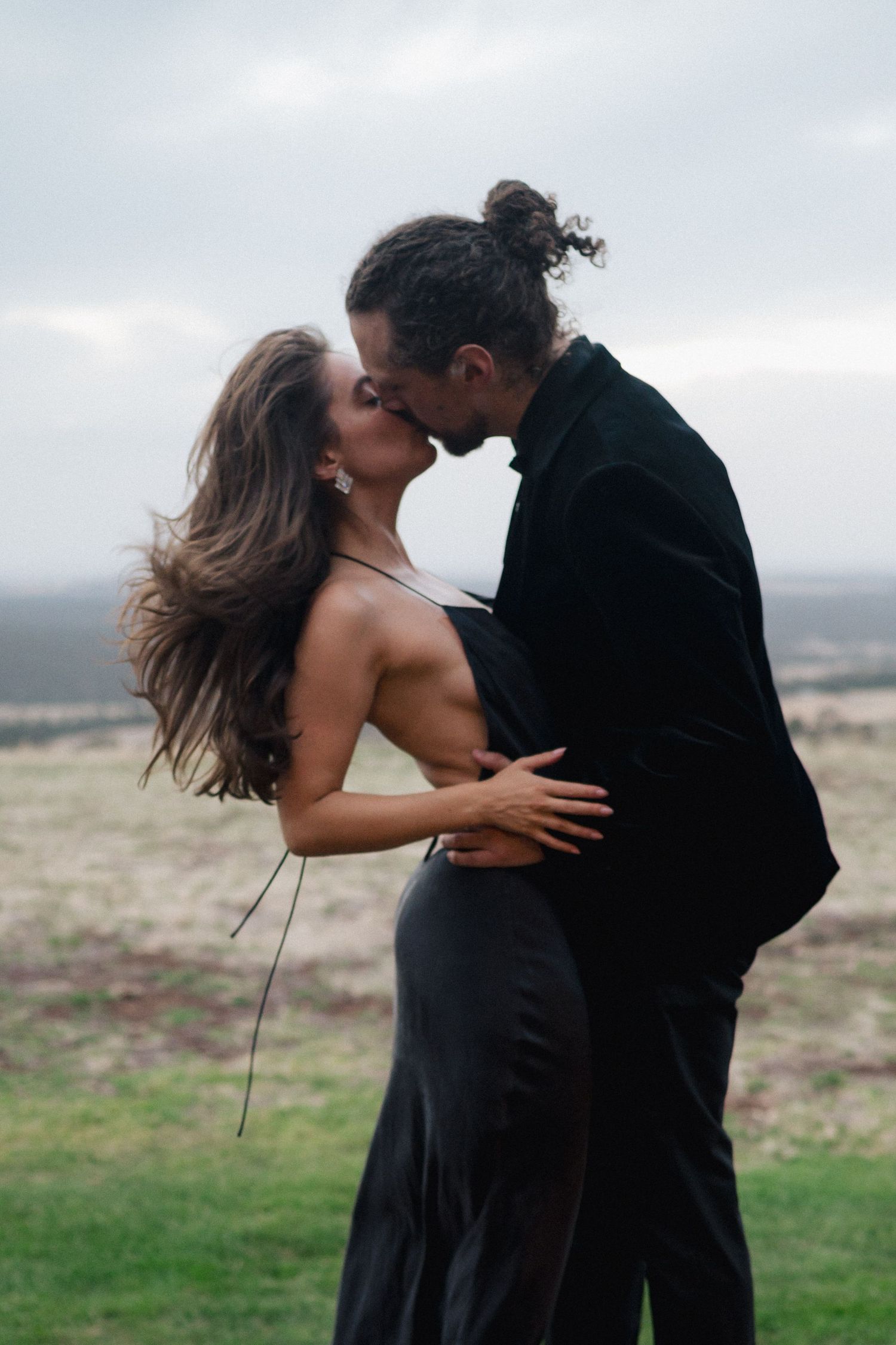 Romantic couple in black attire share intimate moments against a misty outdoor backdrop.