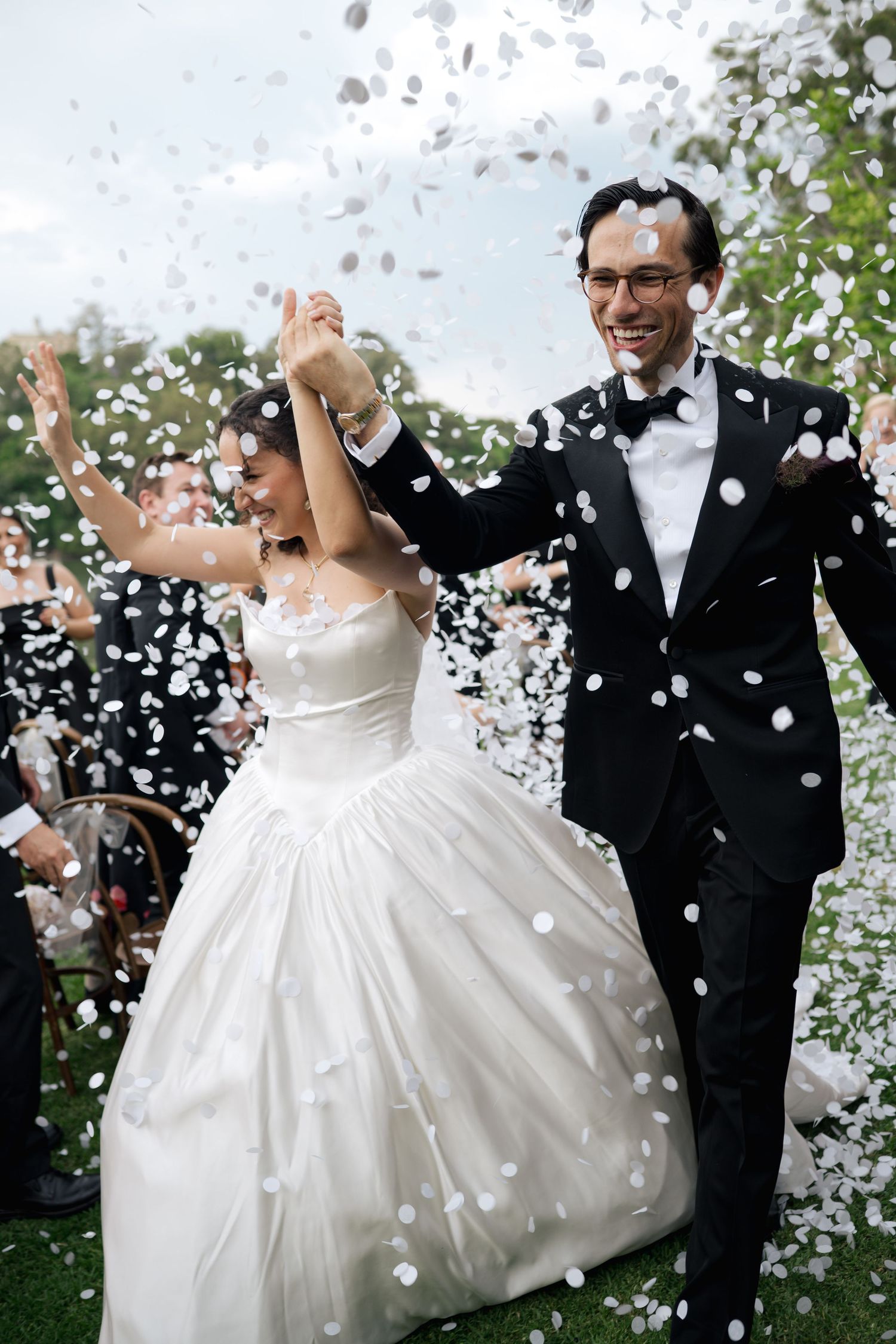 Joyful couple celebrates under a shower of white confetti in an outdoor garden setting.