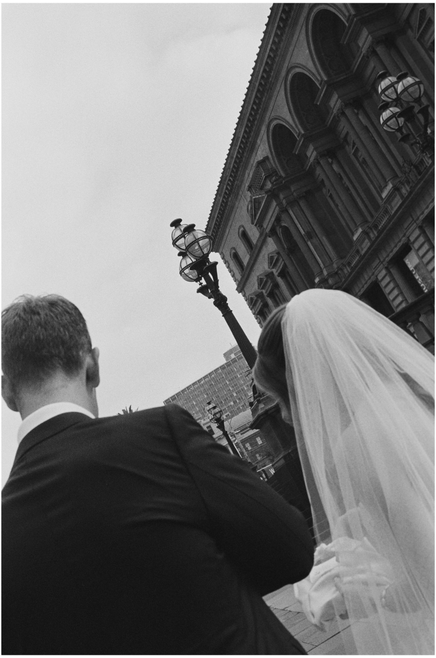 Black and white photo of bride and groom walking toward ornate building, wedding veil flowing, captured from behind.