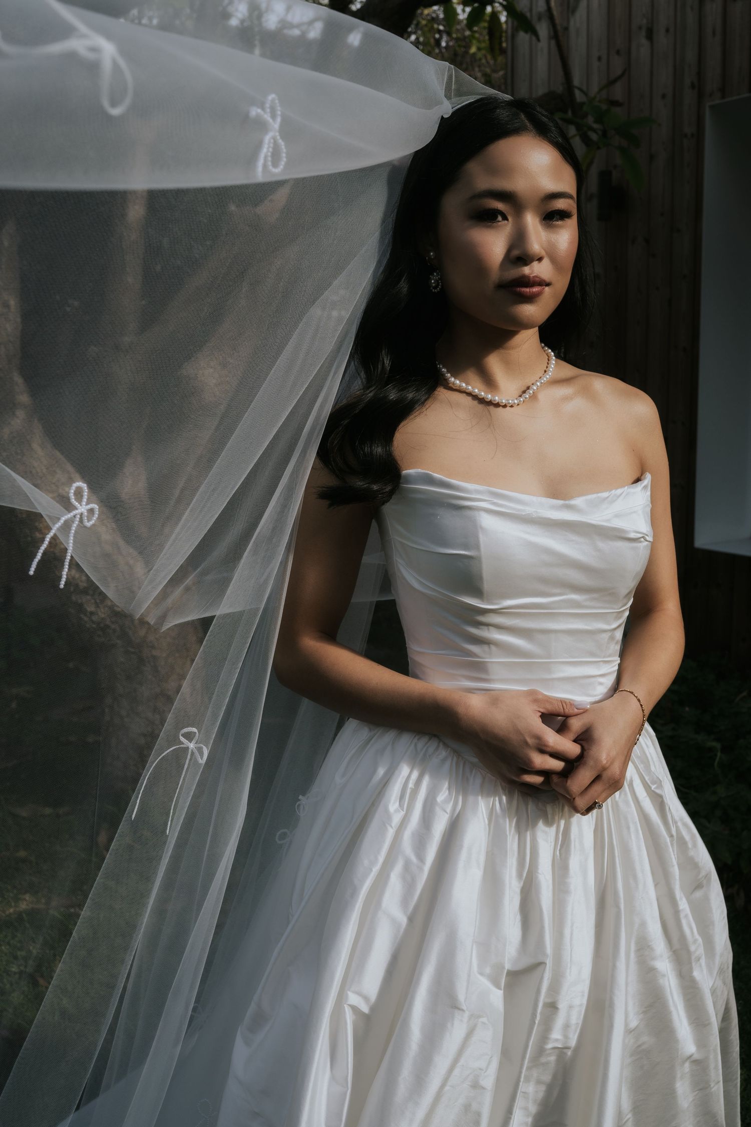 Elegant bride in strapless white ballgown with tulle veil and pearl necklace posing outdoors.