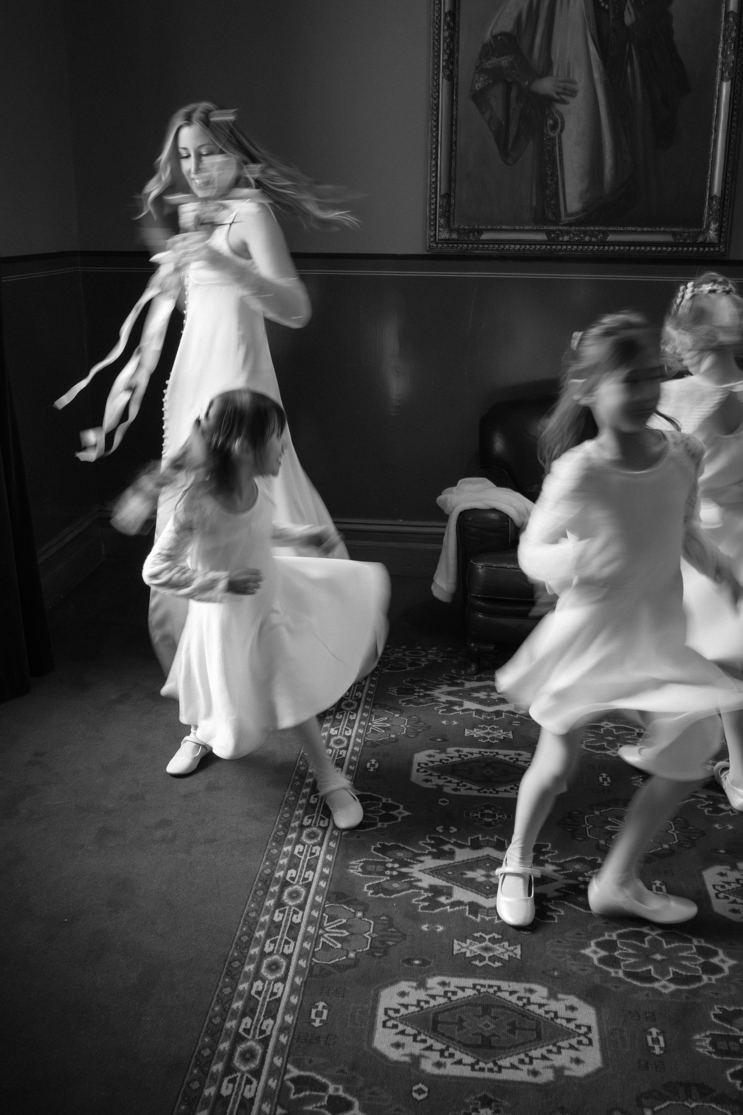 Black and white photo of young girls in white dresses twirling and dancing in an elegant room with a portrait painting.