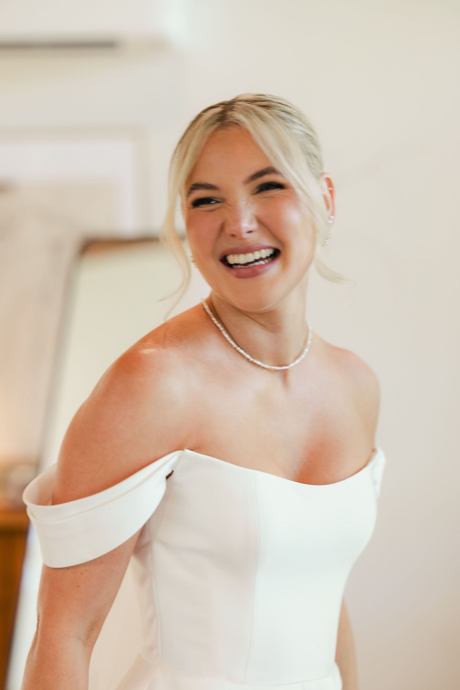 A bride wearing an elegant off-shoulder white wedding gown smiles joyfully during wedding preparations.