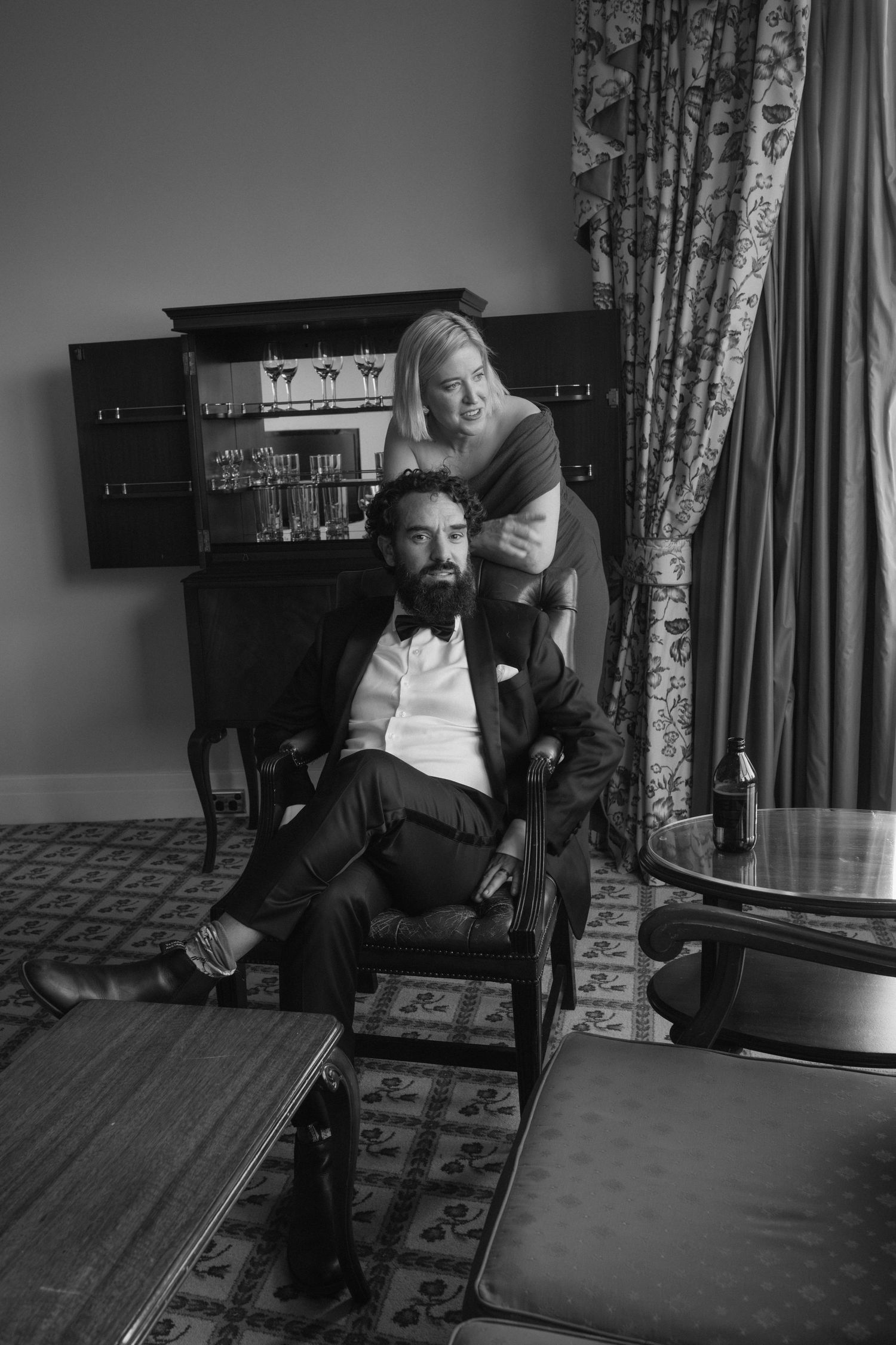 Elegant black and white portrait of a bearded man in tuxedo seated while a woman stands behind him in a sophisticated room.