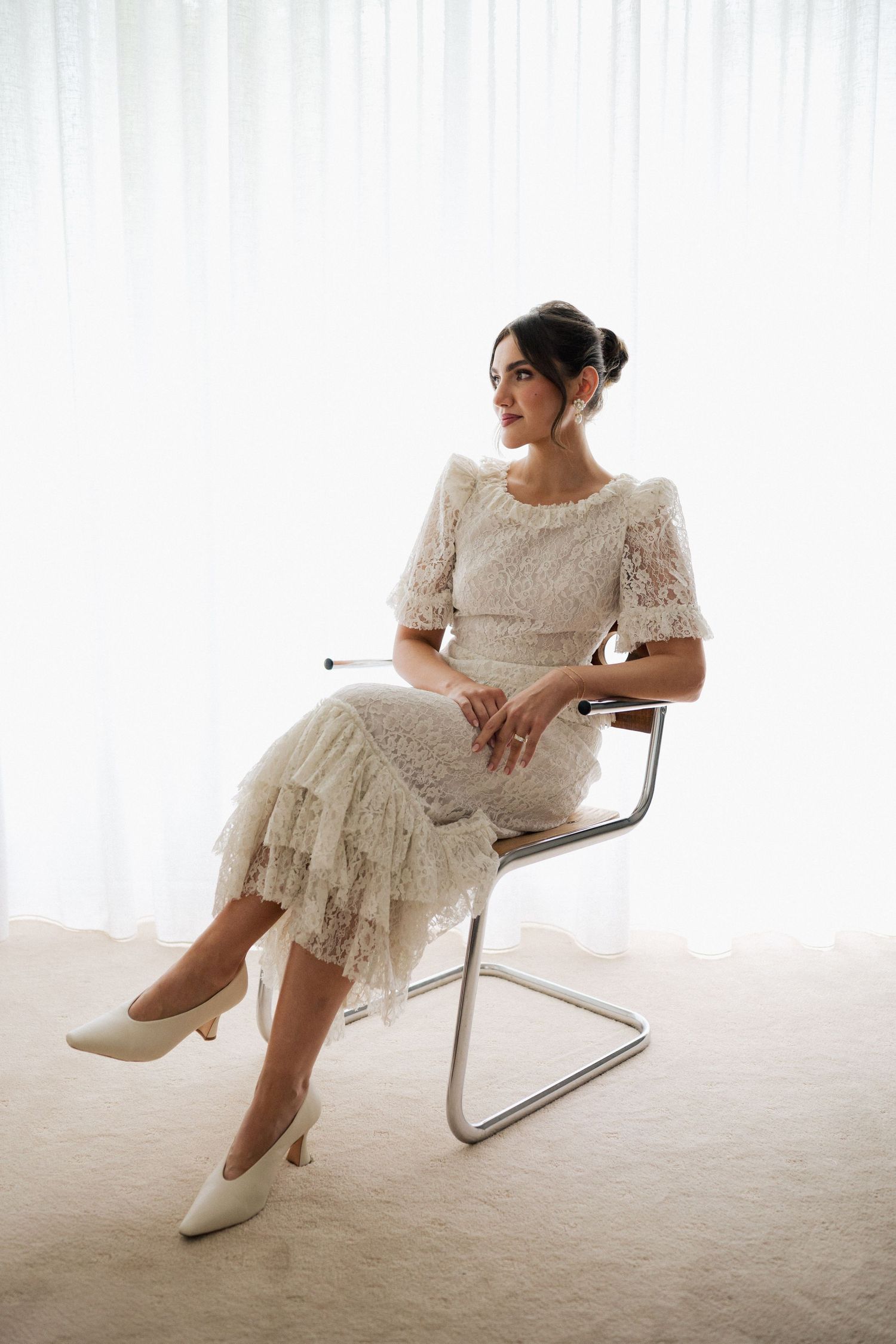 Elegant bride in vintage-style lace dress poses on modern chair in bright studio setting.