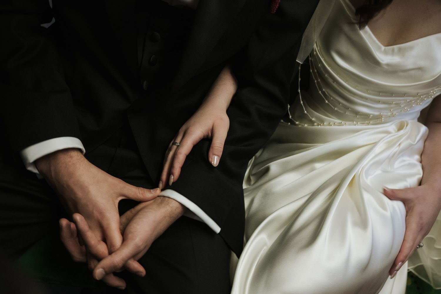 Close-up of bride in white satin gown and groom in black suit holding hands at wedding ceremony.